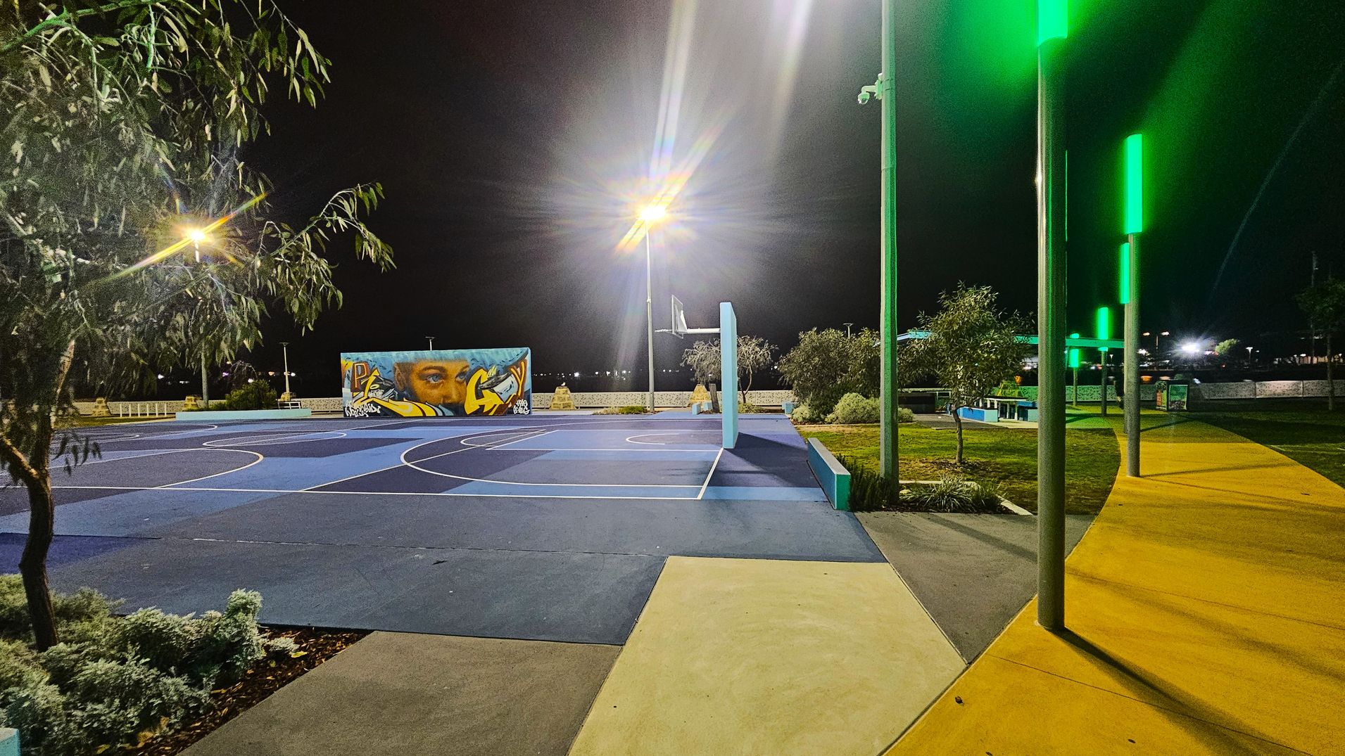 A night-time scene of an outdoor basketball court with a blue surface and white lines. On the left, a tree is visible under a bright streetlight. In the centre, a mural with a face and graffiti is painted on a wall. On the right, a path with yellow paving is lined with tall poles emitting green light. The area is surrounded by small bushes and trees, with additional lighting in the background. A night-time scene of an outdoor basketball court with a blue surface and white lines. On the left, a tree is visible under a bright streetlight. In the centre, a mural with a face and graffiti is painted on a wall. On the right, a path with yellow paving is lined with tall poles emitting green light. The area is surrounded by small bushes and trees, with additional lighting in the background.