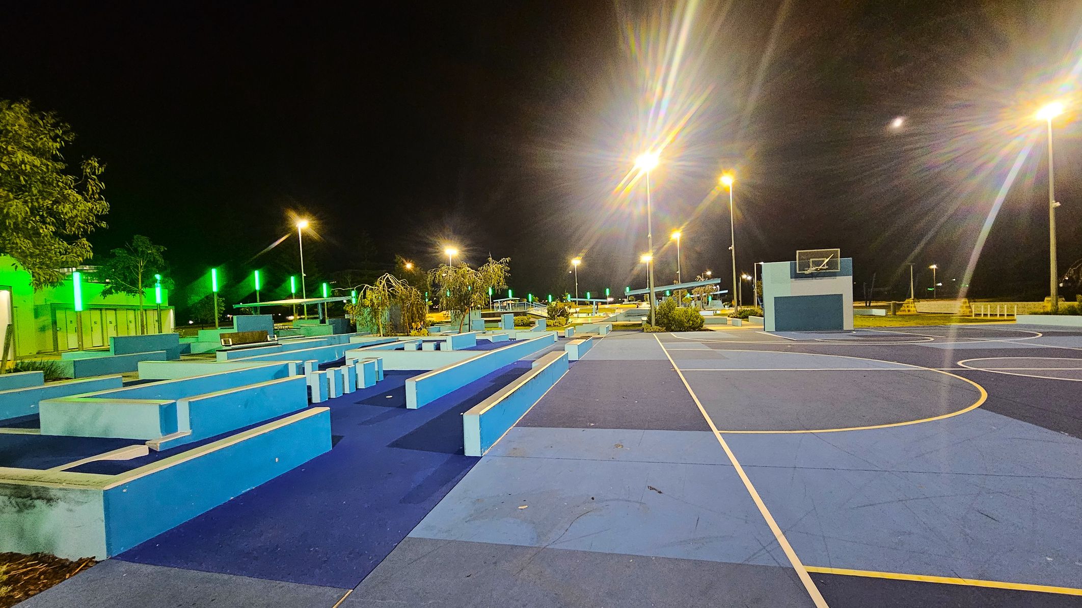 A night-time view of a sports area with a blue and purple basketball court on the right. The court features a hoop with a backboard. On the left, there are geometric blue structures, possibly for seating or parkour. Green lights illuminate the background, with trees and additional structures visible. Tall streetlights cast a warm glow over the scene. The ground is a mix of blue shades, and the sky is dark. A night-time view of a sports area with a blue and purple basketball court on the right. The court features a hoop with a backboard. On the left, there are geometric blue structures, possibly for seating or parkour. Green lights illuminate the background, with trees and additional structures visible. Tall streetlights cast a warm glow over the scene. The ground is a mix of blue shades, and the sky is dark.