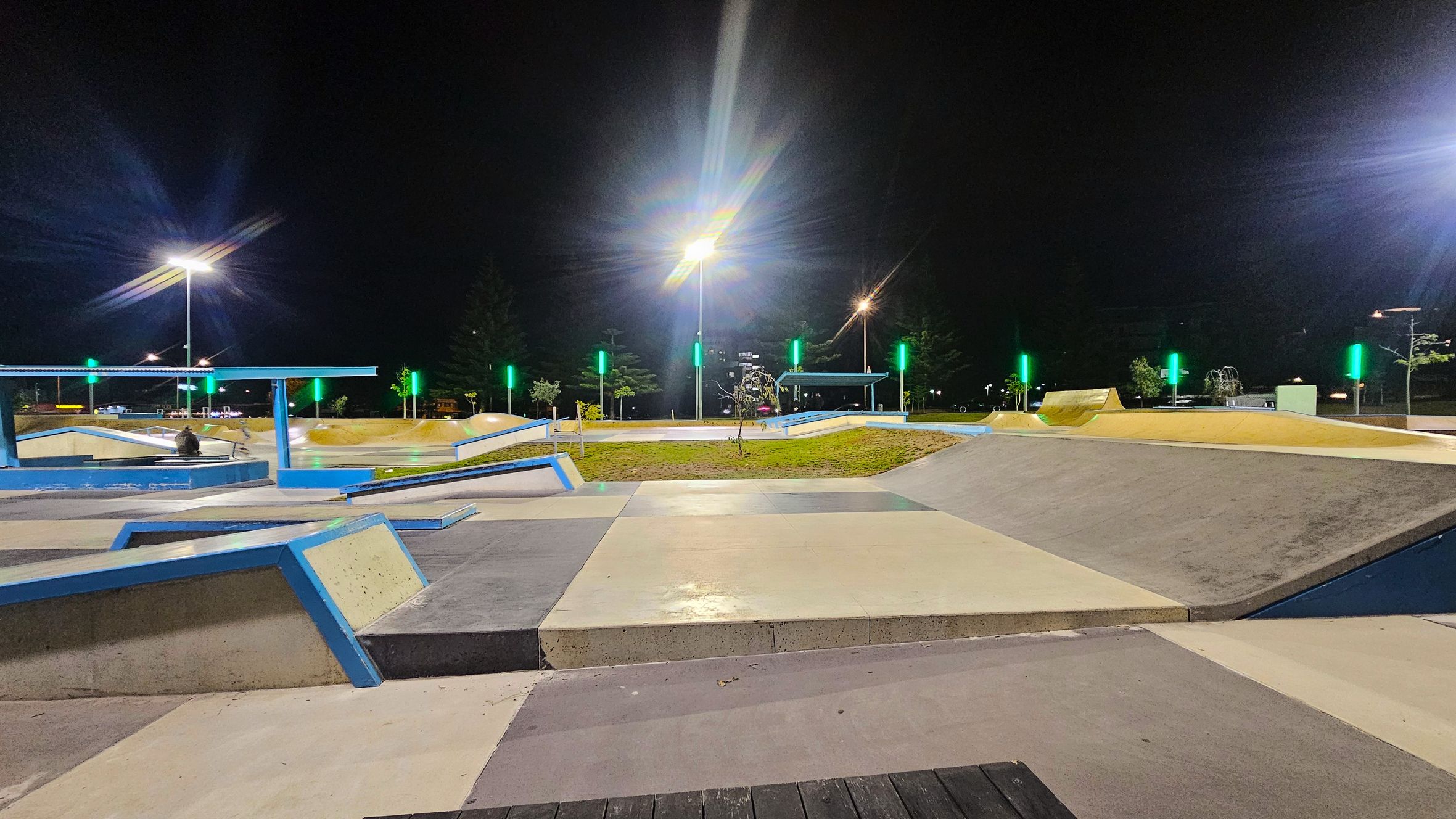 A well-lit skate park at night with various ramps and platforms. The foreground features a smooth concrete surface with blue-edged ramps. In the middle, there are grassy areas and more ramps, with several tall lights illuminating the scene. The background shows trees and additional park structures, with green lights accentuating the area. The sky is dark, indicating nighttime. A well-lit skate park at night with various ramps and platforms. The foreground features a smooth concrete surface with blue-edged ramps. In the middle, there are grassy areas and more ramps, with several tall lights illuminating the scene. The background shows trees and additional park structures, with green lights accentuating the area. The sky is dark, indicating nighttime.