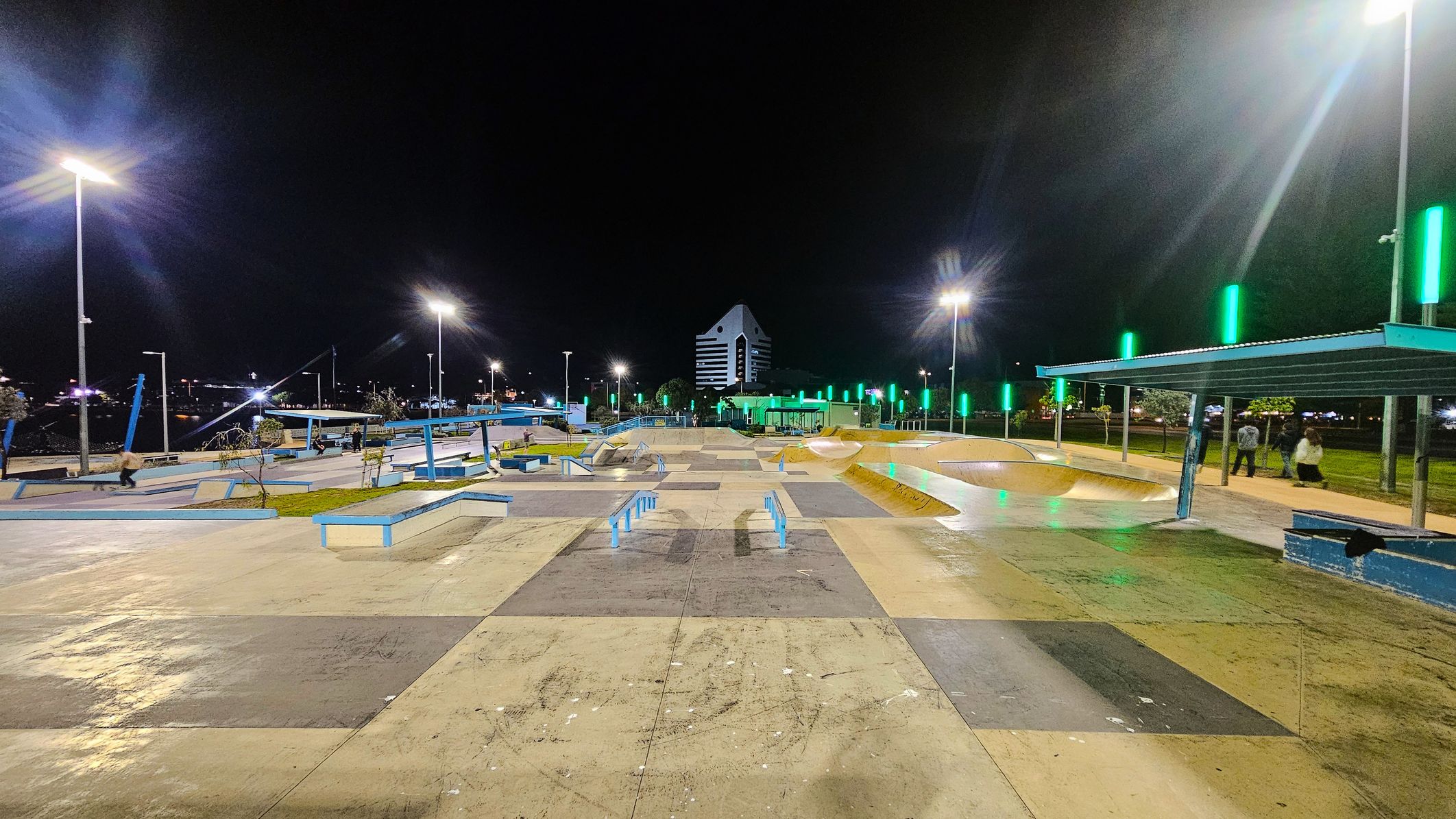A well-lit skate park at night with various ramps and rails. The ground is a mix of concrete and coloured sections. On the right, there's a covered seating area with people standing nearby. Green lights line the path, and tall streetlights illuminate the area. In the background, a distinctive building with a triangular roof and striped facade is visible. The park is surrounded by trees and additional lighting. A well-lit skate park at night with various ramps and rails. The ground is a mix of concrete and coloured sections. On the right, there's a covered seating area with people standing nearby. Green lights line the path, and tall streetlights illuminate the area. In the background, a distinctive building with a triangular roof and striped facade is visible. The park is surrounded by trees and additional lighting.