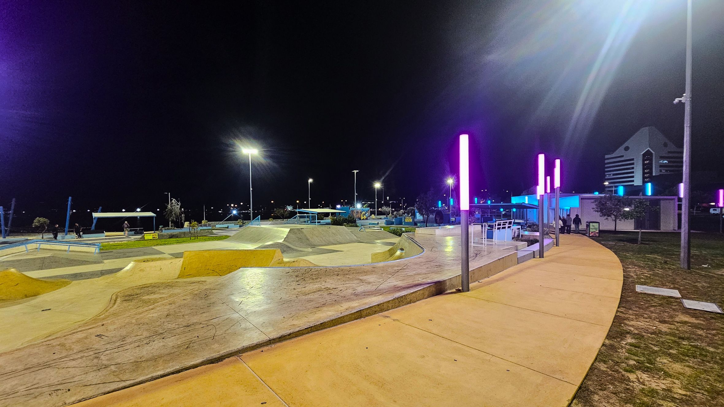 A skate park at night with various ramps and rails on the left. The ground is a mix of concrete and grass. On the right, a curved pathway is lined with tall poles featuring pink and blue lights. In the background, a modern building with a triangular roof is visible. The area is well-lit with streetlights, and a few people are present near the structures. The sky is dark, indicating nighttime. A skate park at night with various ramps and rails on the left. The ground is a mix of concrete and grass. On the right, a curved pathway is lined with tall poles featuring pink and blue lights. In the background, a modern building with a triangular roof is visible. The area is well-lit with streetlights, and a few people are present near the structures. The sky is dark, indicating nighttime.