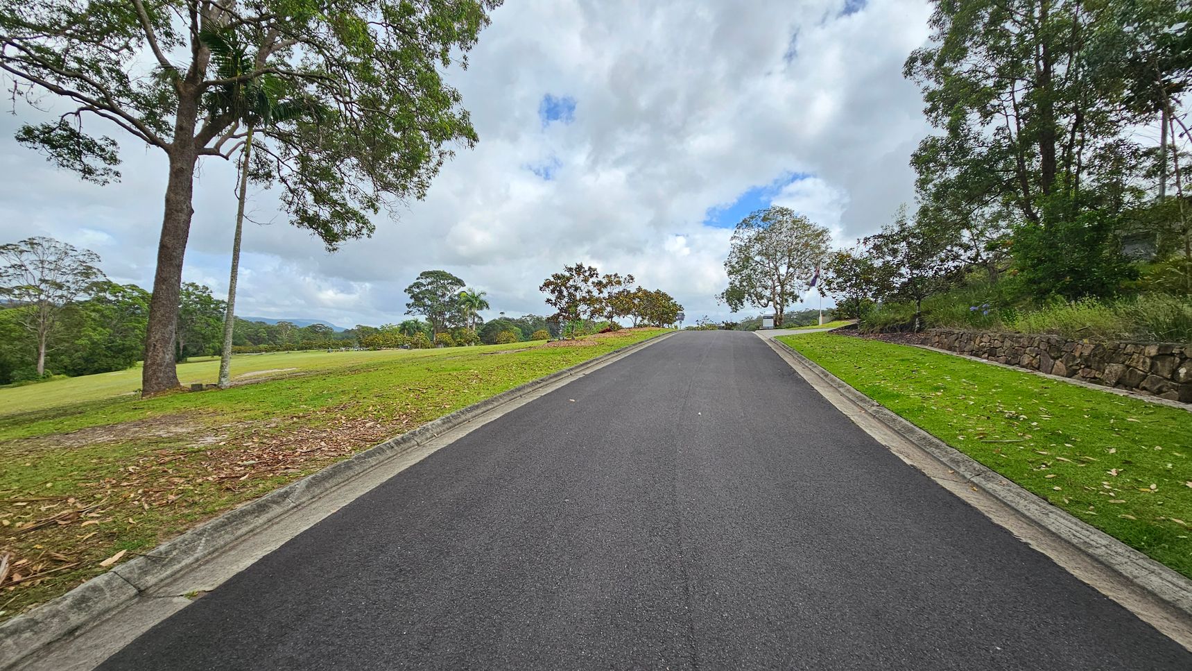 A paved road leads uphill, flanked by tall trees and greenery on both sides. On the left, a sign reads "Kulangoor Cemetery Est. 1907" near a large tree. On the right, a small metal box is attached to a concrete pillar. The road is bordered by grass and shrubs, with speed limit signs visible. The sky is overcast, and the area is surrounded by dense foliage. A paved road leads uphill, flanked by tall trees and greenery on both sides. On the left, a sign reads "Kulangoor Cemetery Est. 1907" near a large tree. On the right, a small metal box is attached to a concrete pillar. The road is bordered by grass and shrubs, with speed limit signs visible. The sky is overcast, and the area is surrounded by dense foliage.