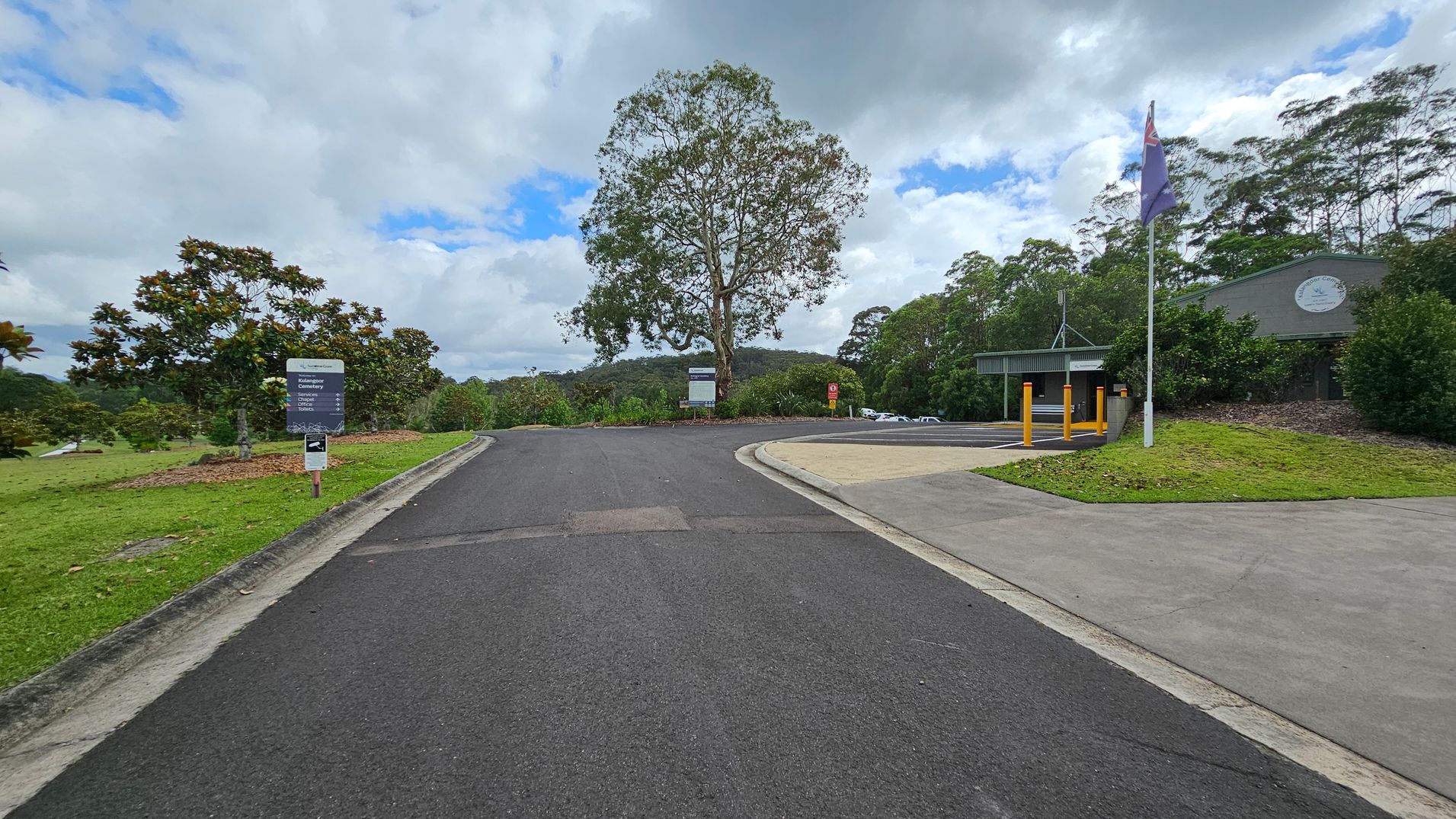 A paved road leads to a T-junction with a large tree in the centre. On the left, there's a grassy area with several smaller trees and a sign. On the right, a building with a green roof and a flagpole stands near a parking area. The background features a forested hill under a cloudy sky. A paved road leads to a T-junction with a large tree in the centre. On the left, there's a grassy area with several smaller trees and a sign. On the right, a building with a green roof and a flagpole stands near a parking area. The background features a forested hill under a cloudy sky.
