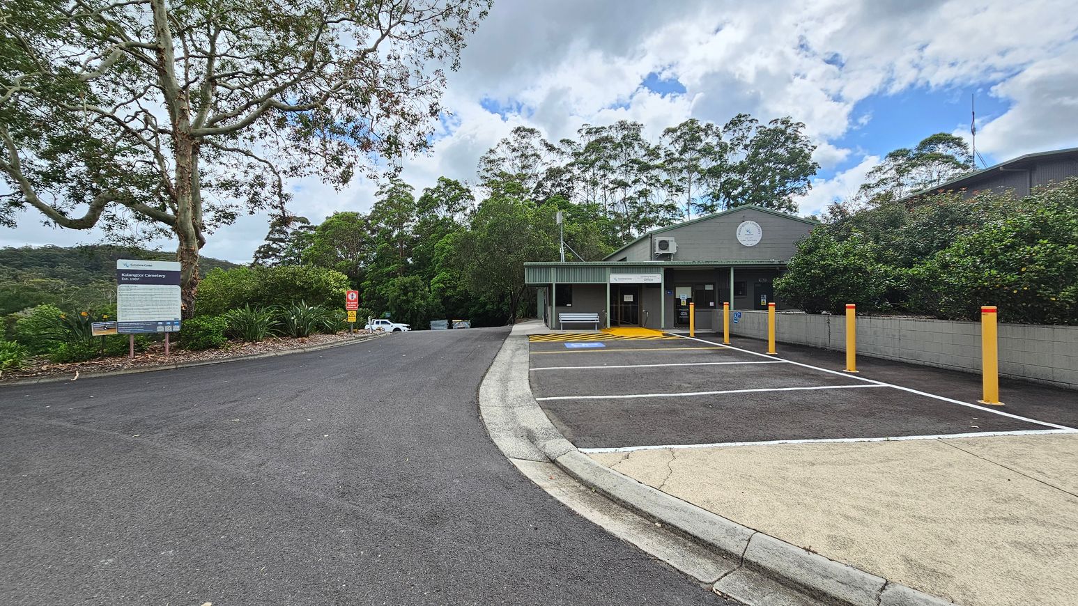 A paved road leads to a building on the right with a green roof and a clock on the facade. Yellow bollards line the edge of the parking area, which includes a disabled parking space. On the left, a large tree stands next to a sign with text, surrounded by greenery. In the background, dense trees cover the hillside. A white vehicle is partially visible further down the road. A paved road leads to a building on the right with a green roof and a clock on the facade. Yellow bollards line the edge of the parking area, which includes a disabled parking space. On the left, a large tree stands next to a sign with text, surrounded by greenery. In the background, dense trees cover the hillside. A white vehicle is partially visible further down the road.