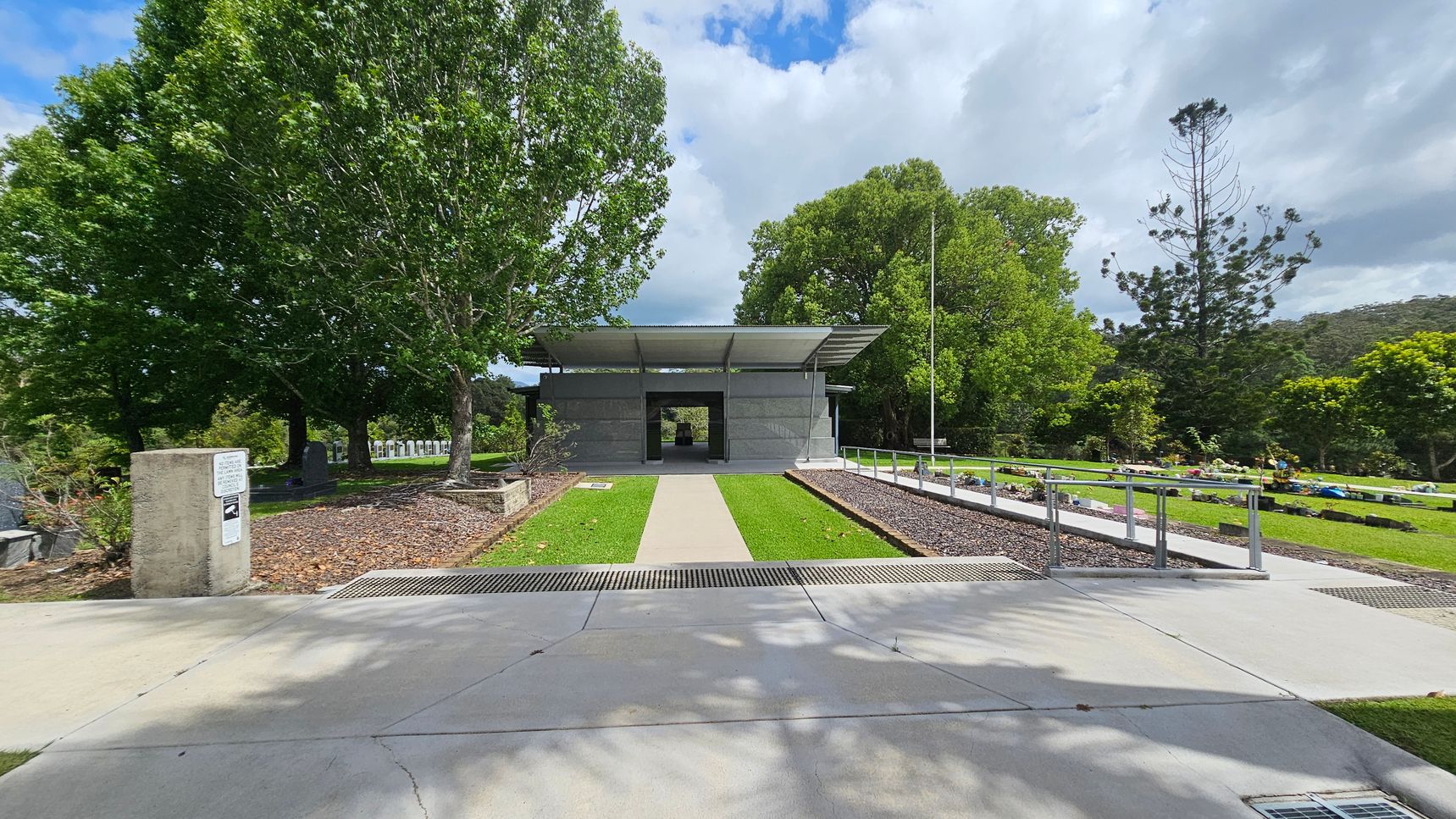 A pathway leads to a modern, open-sided structure with a flat roof, surrounded by lush green trees. On the left, a concrete post with signage stands near a tree-lined area with gravestones. On the right, a manicured lawn with a row of low plants and a metal railing. The background features more trees and a slightly overcast sky. The foreground shows a concrete pavement with shadows from the trees. A pathway leads to a modern, open-sided structure with a flat roof, surrounded by lush green trees. On the left, a concrete post with signage stands near a tree-lined area with gravestones. On the right, a manicured lawn with a row of low plants and a metal railing. The background features more trees and a slightly overcast sky. The foreground shows a concrete pavement with shadows from the trees.