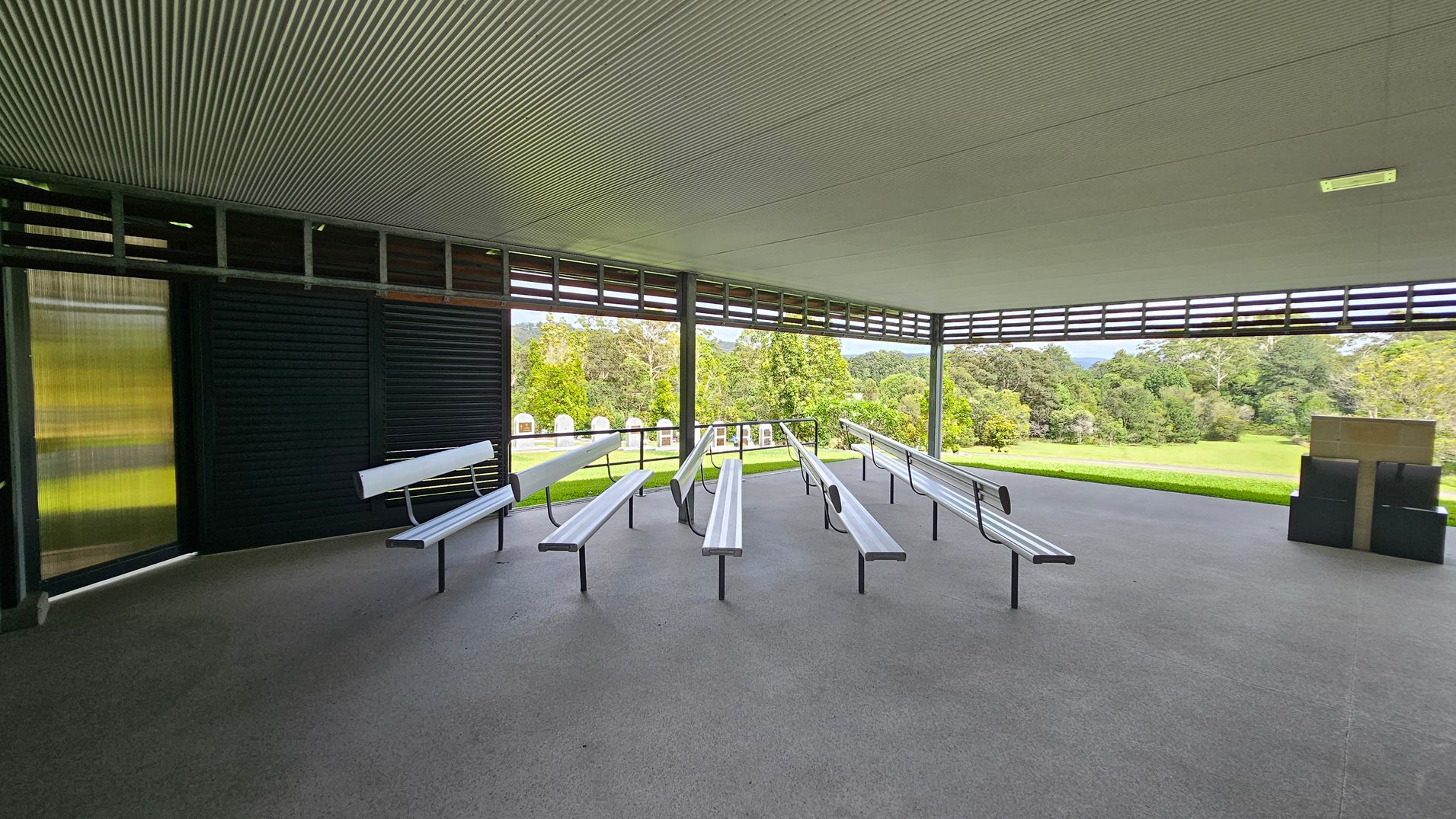 An open-air pavilion with a corrugated ceiling and grey floor. In the centre, there are several rows of metal benches facing forward. The left side has a dark louvred wall, while the right side opens to a grassy area with trees in the background. A stack of black boxes is on the right. The pavilion is surrounded by a lush, green landscape. An open-air pavilion with a corrugated ceiling and grey floor. In the centre, there are several rows of metal benches facing forward. The left side has a dark louvred wall, while the right side opens to a grassy area with trees in the background. A stack of black boxes is on the right. The pavilion is surrounded by a lush, green landscape.