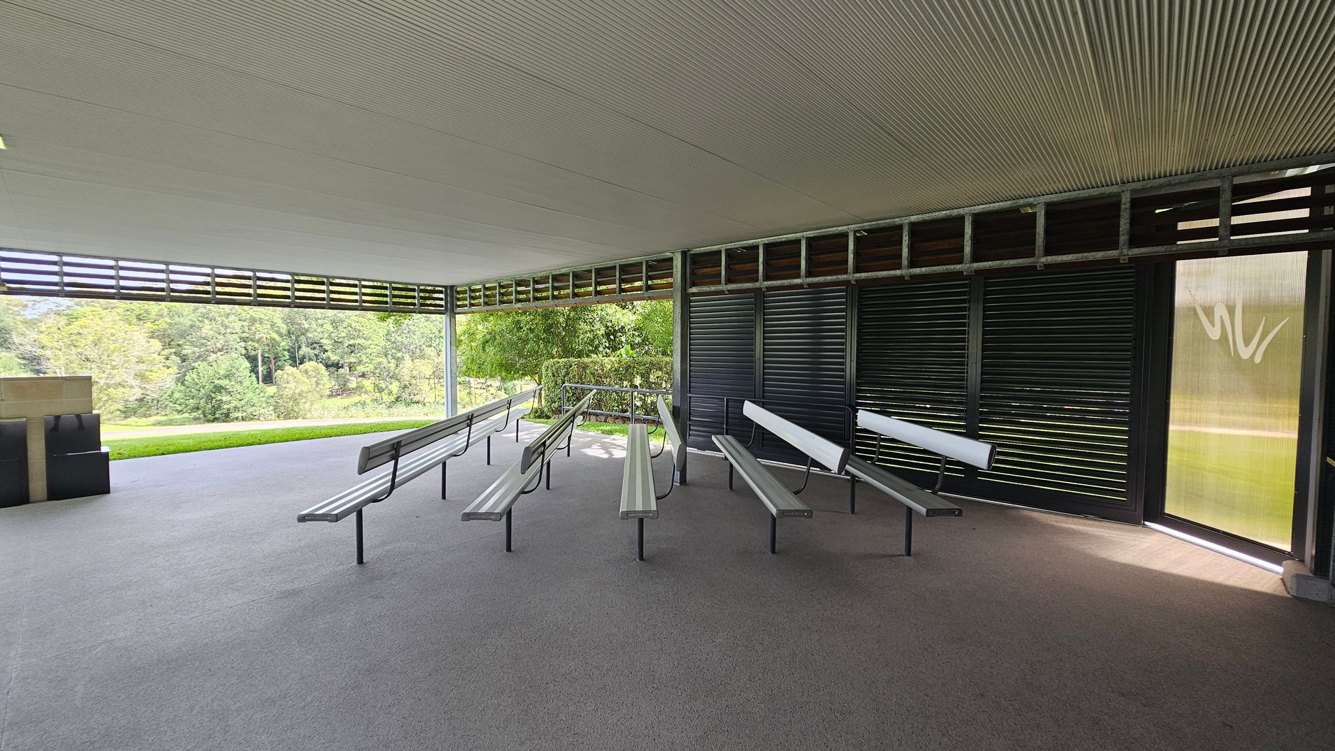 An open-air pavilion with a grey, textured floor and a ribbed ceiling. In the centre, there are several metal benches with backrests arranged in rows. The right side features dark louvred panels, while the left side is open, offering a view of lush green trees and grass. A small structure with a flat top is visible on the far left. Natural light filters through the space, creating a serene atmosphere. An open-air pavilion with a grey, textured floor and a ribbed ceiling. In the centre, there are several metal benches with backrests arranged in rows. The right side features dark louvred panels, while the left side is open, offering a view of lush green trees and grass. A small structure with a flat top is visible on the far left. Natural light filters through the space, creating a serene atmosphere.