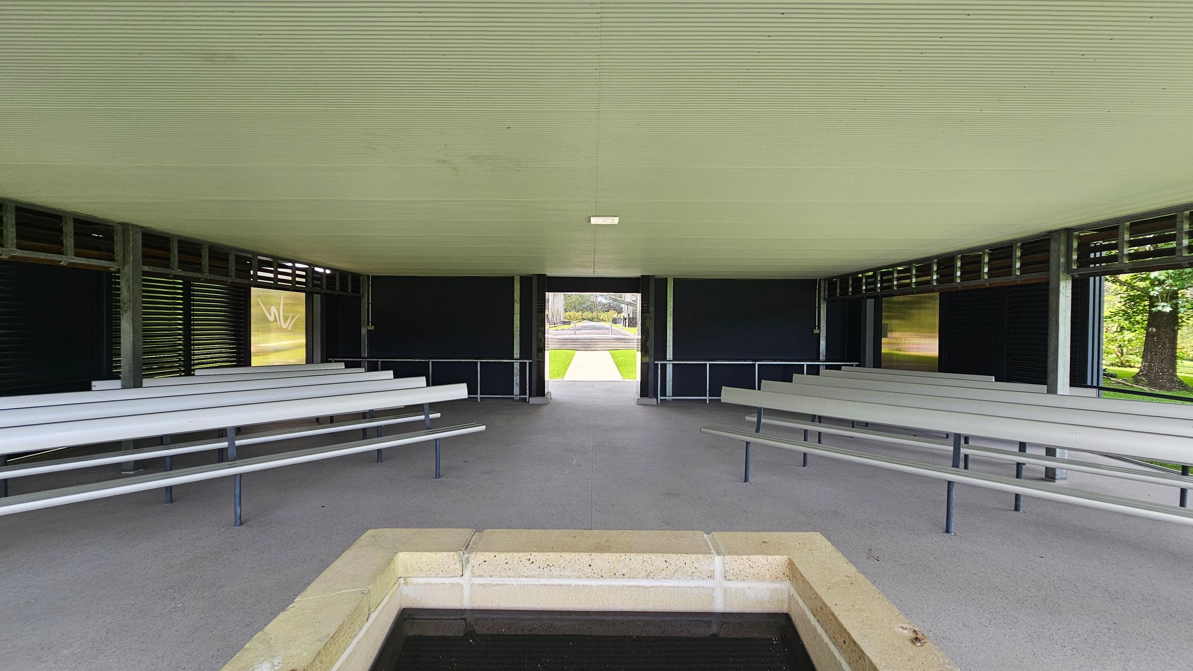 A covered outdoor pavilion with a corrugated ceiling and concrete floor. In the foreground, there is a rectangular stone structure, possibly a water feature. On both sides, there are rows of grey benches facing inward. The background shows an open exit leading to a grassy area and a path. The walls have dark panels with horizontal slats, allowing some light to filter through. Trees are visible outside on the right. A covered outdoor pavilion with a corrugated ceiling and concrete floor. In the foreground, there is a rectangular stone structure, possibly a water feature. On both sides, there are rows of grey benches facing inward. The background shows an open exit leading to a grassy area and a path. The walls have dark panels with horizontal slats, allowing some light to filter through. Trees are visible outside on the right.