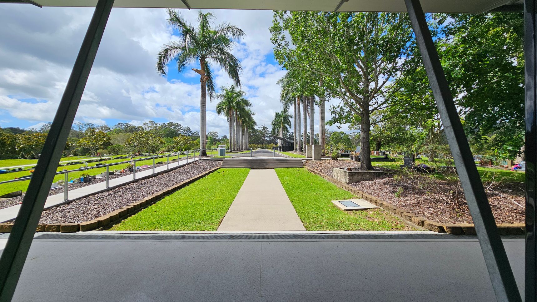 A pathway leads through a garden with neatly trimmed grass, flanked by tall palm trees on both sides. On the left, a railing runs alongside a gravel path, with more greenery and distant trees. On the right, a large tree stands in a raised bed with mulch. In the background, a structure is partially visible, surrounded by more trees. The foreground shows a covered area with a grey floor and metal beams. A pathway leads through a garden with neatly trimmed grass, flanked by tall palm trees on both sides. On the left, a railing runs alongside a gravel path, with more greenery and distant trees. On the right, a large tree stands in a raised bed with mulch. In the background, a structure is partially visible, surrounded by more trees. The foreground shows a covered area with a grey floor and metal beams.