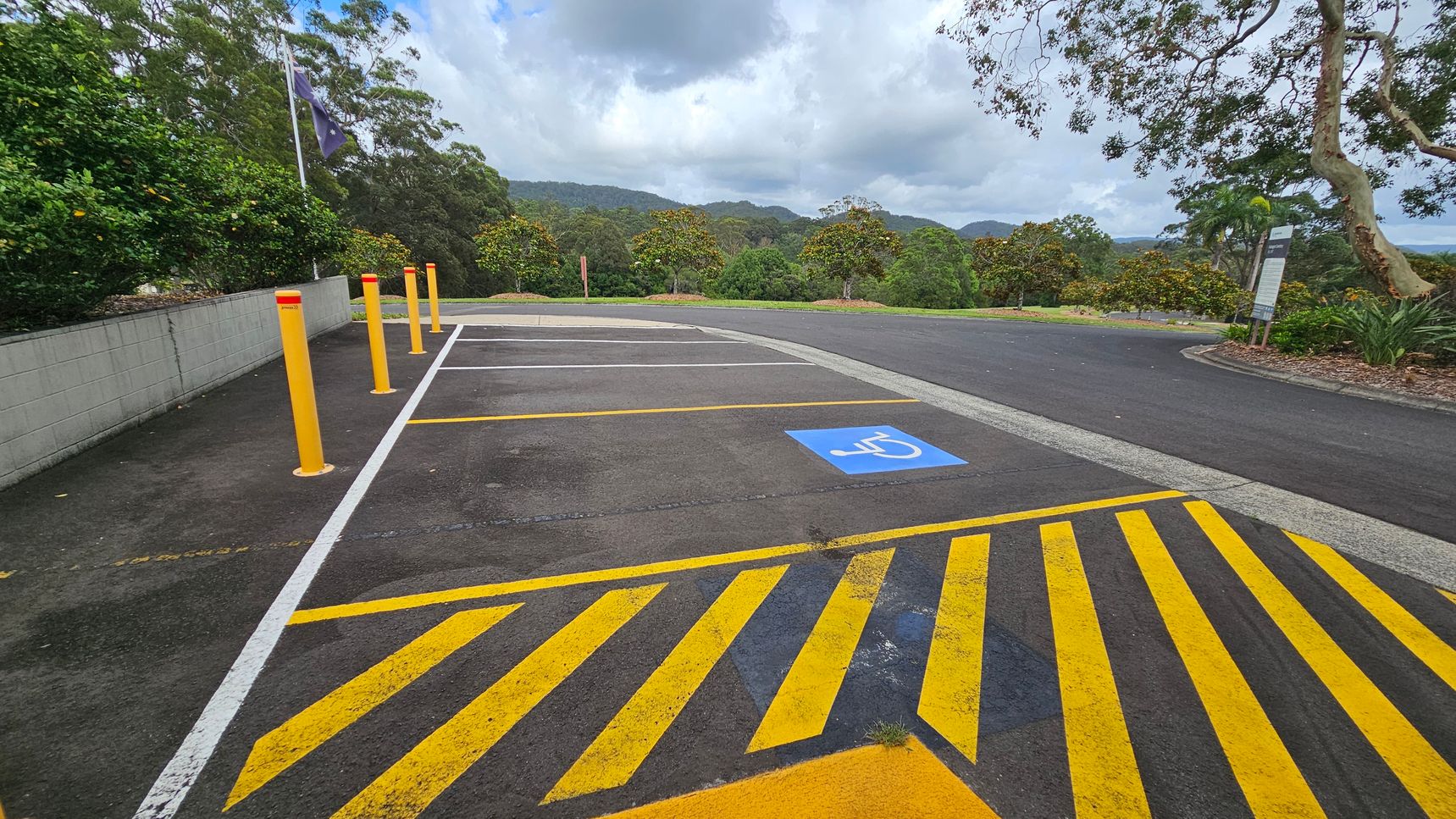 A parking area with a designated disabled parking space marked by a blue symbol on the ground. Yellow diagonal lines are painted on the asphalt in the foreground. On the left, there are several yellow bollards and a low concrete wall with greenery behind it. The background features lush trees and hills under a cloudy sky. On the right, a sign is visible near more vegetation. The area is surrounded by natural scenery. A parking area with a designated disabled parking space marked by a blue symbol on the ground. Yellow diagonal lines are painted on the asphalt in the foreground. On the left, there are several yellow bollards and a low concrete wall with greenery behind it. The background features lush trees and hills under a cloudy sky. On the right, a sign is visible near more vegetation. The area is surrounded by natural scenery.