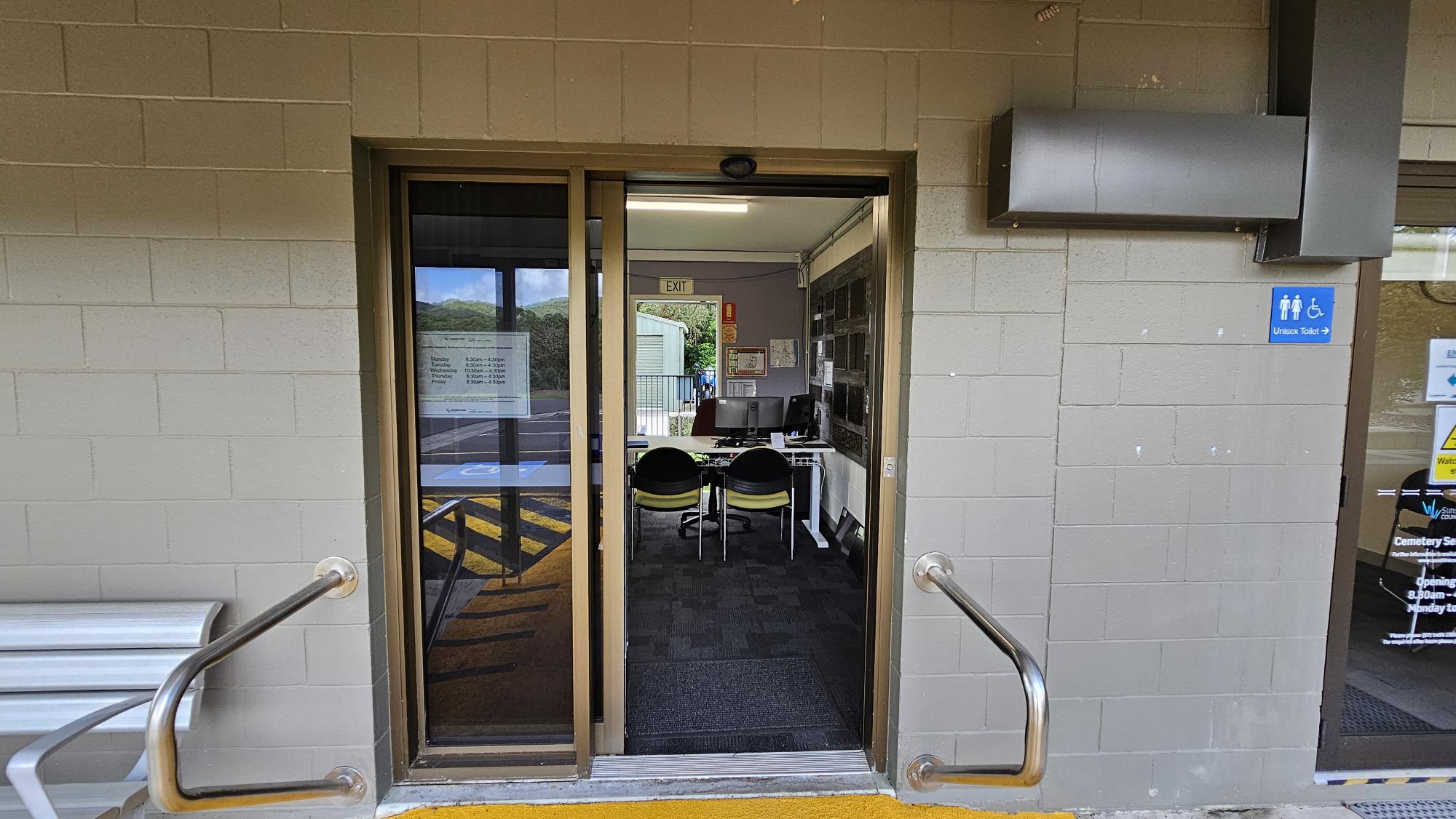 A doorway with a sliding glass door leads into an office space. Inside, there are two chairs facing a desk with a computer. The walls are made of light-coloured bricks. On the right, a sign indicates the direction to a unisex toilet. Metal handrails are on either side of the entrance. The floor outside is marked with yellow paint, and the interior has dark carpeting. An "EXIT" sign is visible inside above the door. A doorway with a sliding glass door leads into an office space. Inside, there are two chairs facing a desk with a computer. The walls are made of light-coloured bricks. On the right, a sign indicates the direction to a unisex toilet. Metal handrails are on either side of the entrance. The floor outside is marked with yellow paint, and the interior has dark carpeting. An "EXIT" sign is visible inside above the door.