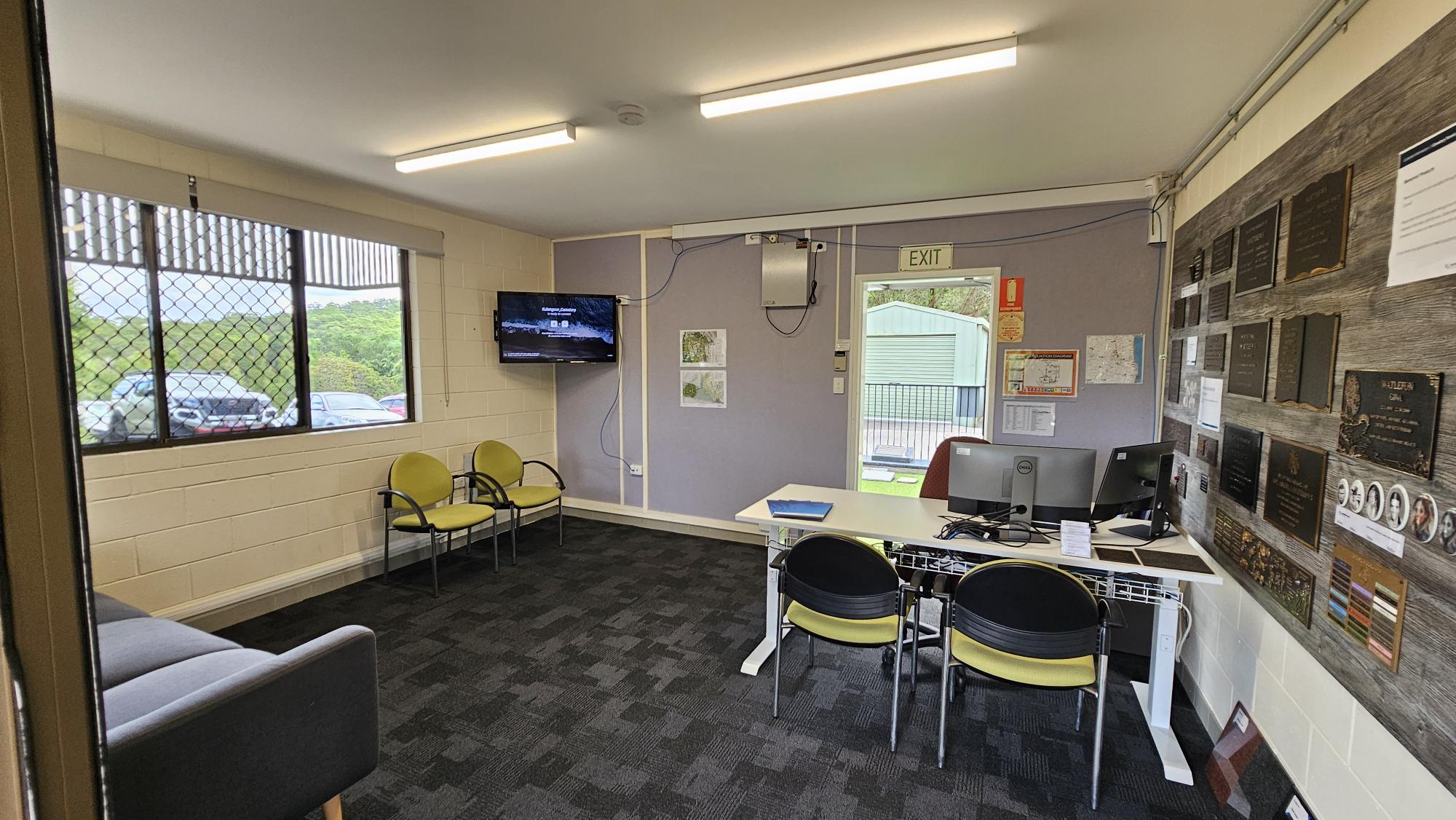 A room with a dark carpeted floor and fluorescent lights on the ceiling. On the left, there are windows with metal grilles and a view of parked cars outside. Below the windows, a grey sofa and two yellow chairs are placed. In the middle, a wall-mounted TV is visible. On the right, a desk with two computer monitors and chairs is positioned. The wall behind the desk is covered with plaques and notices. An open door leads outside, showing a green building in the background. A room with a dark carpeted floor and fluorescent lights on the ceiling. On the left, there are windows with metal grilles and a view of parked cars outside. Below the windows, a grey sofa and two yellow chairs are placed. In the middle, a wall-mounted TV is visible. On the right, a desk with two computer monitors and chairs is positioned. The wall behind the desk is covered with plaques and notices. An open door leads outside, showing a green building in the background.