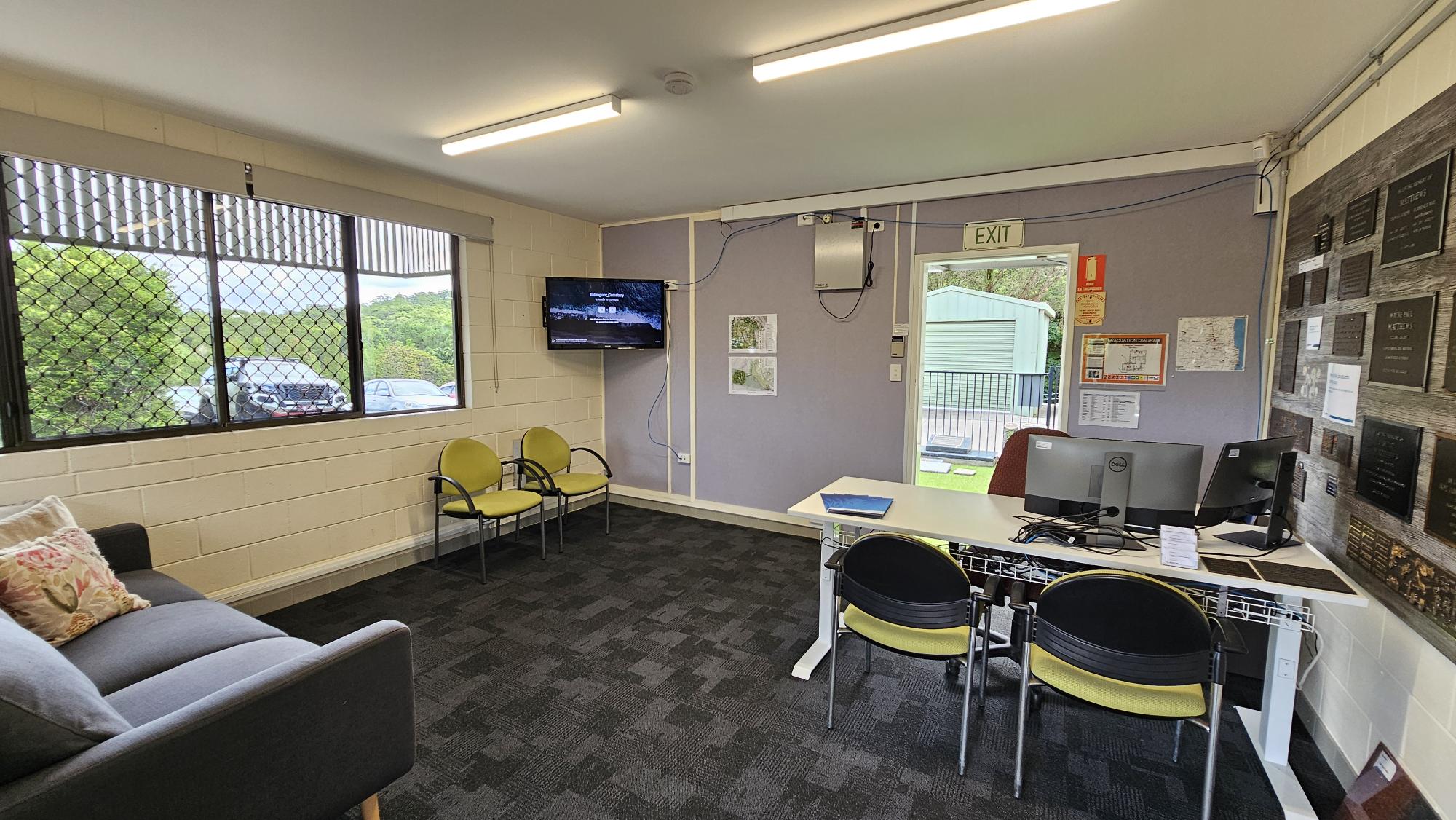 A small office space with a grey sofa and colourful cushions on the left, next to a large window with a metal grid. Two yellow chairs are positioned near the window. In the middle, a wall-mounted TV is visible. On the right, a desk with two computer monitors and chairs faces a wall adorned with plaques. An exit door is nearby, leading outside. The floor is carpeted in a dark pattern, and the ceiling has fluorescent lights. A small office space with a grey sofa and colourful cushions on the left, next to a large window with a metal grid. Two yellow chairs are positioned near the window. In the middle, a wall-mounted TV is visible. On the right, a desk with two computer monitors and chairs faces a wall adorned with plaques. An exit door is nearby, leading outside. The floor is carpeted in a dark pattern, and the ceiling has fluorescent lights.