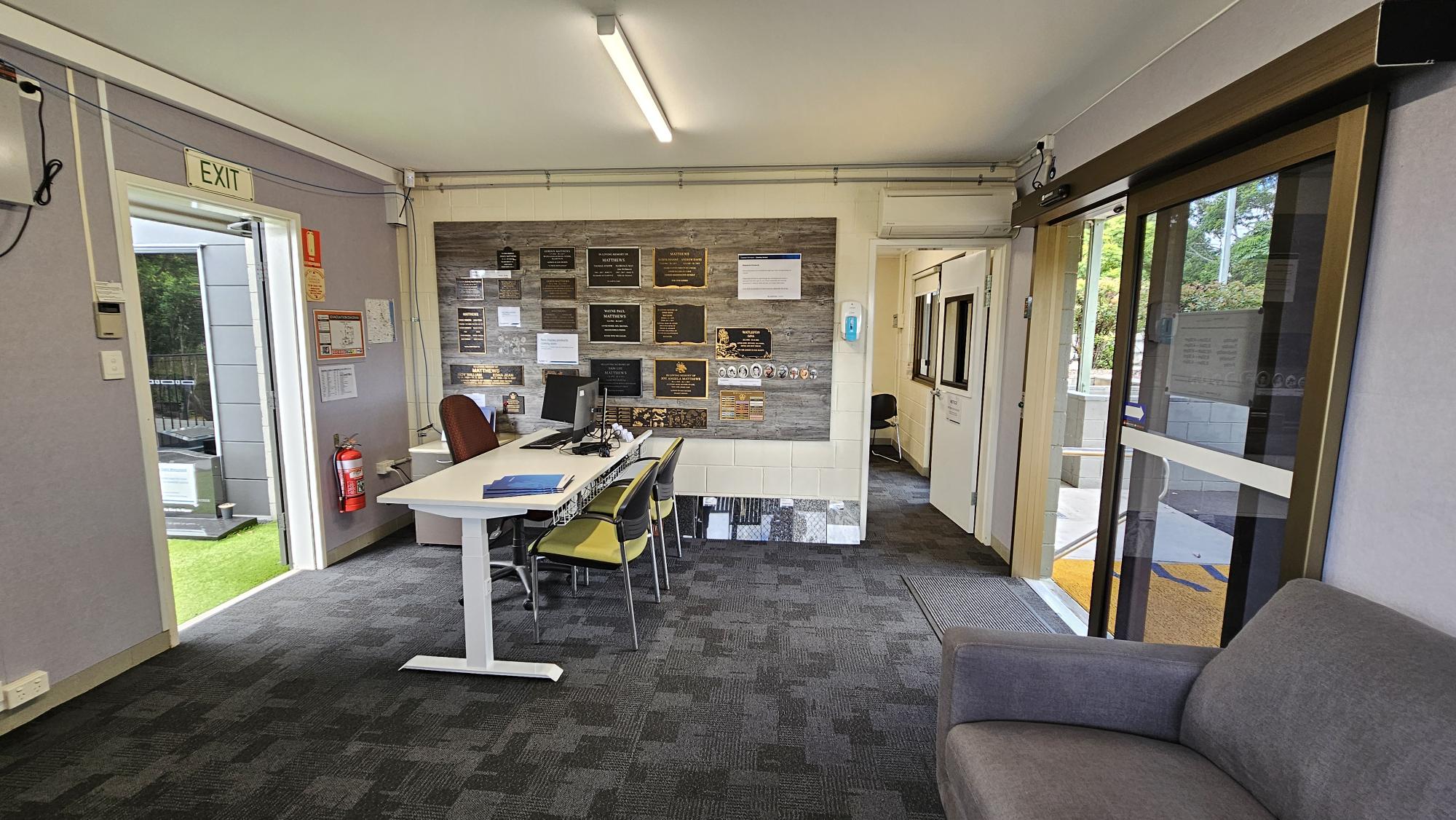 A small office space with a grey carpeted floor. On the left, there's an open door with an "EXIT" sign above it and a fire extinguisher nearby. In the centre, a white desk with a computer and chairs faces a wall adorned with various plaques and notices. On the right, a glass sliding door leads outside, and a grey sofa is partially visible. The ceiling has a fluorescent light fixture. A small office space with a grey carpeted floor. On the left, there's an open door with an "EXIT" sign above it and a fire extinguisher nearby. In the centre, a white desk with a computer and chairs faces a wall adorned with various plaques and notices. On the right, a glass sliding door leads outside, and a grey sofa is partially visible. The ceiling has a fluorescent light fixture.