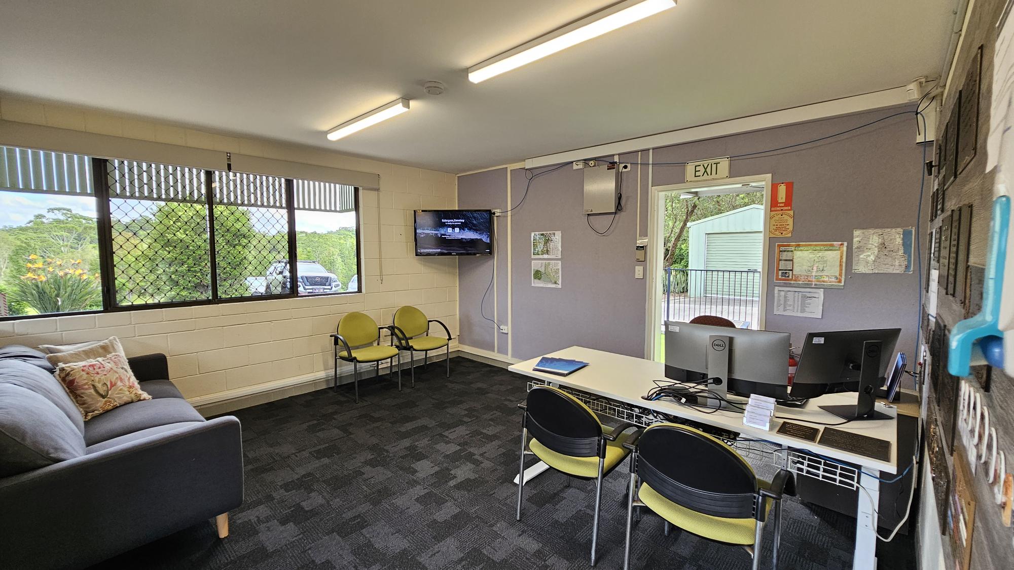 A room with a grey sofa and decorative cushions on the left, next to a window with a view of greenery and a parked car outside. In the middle, there are two yellow chairs and a wall-mounted TV. On the right, a desk with two computer monitors and chairs. The wall has maps and an exit sign above a door leading outside. The floor is carpeted in dark grey, and the ceiling has fluorescent lights. A room with a grey sofa and decorative cushions on the left, next to a window with a view of greenery and a parked car outside. In the middle, there are two yellow chairs and a wall-mounted TV. On the right, a desk with two computer monitors and chairs. The wall has maps and an exit sign above a door leading outside. The floor is carpeted in dark grey, and the ceiling has fluorescent lights.