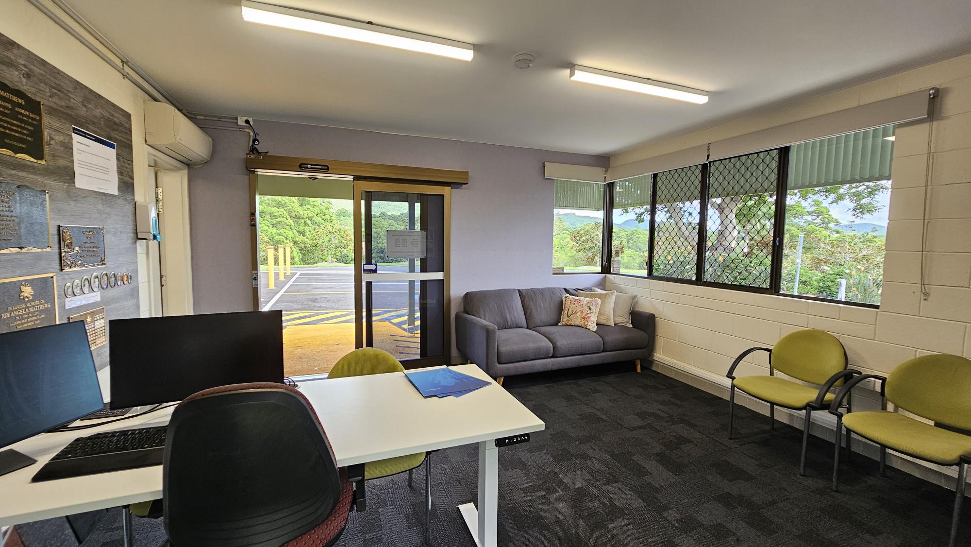 A small office space with a desk on the left featuring two computer monitors and a chair. The wall behind the desk has several plaques and notices. In the middle, a glass door leads outside, allowing natural light in. On the right, a grey sofa with decorative cushions is placed against a wall with large windows, offering a view of greenery outside. Two green chairs are positioned near the sofa. The floor is carpeted, and the ceiling has fluorescent lights. A small office space with a desk on the left featuring two computer monitors and a chair. The wall behind the desk has several plaques and notices. In the middle, a glass door leads outside, allowing natural light in. On the right, a grey sofa with decorative cushions is placed against a wall with large windows, offering a view of greenery outside. Two green chairs are positioned near the sofa. The floor is carpeted, and the ceiling has fluorescent lights.