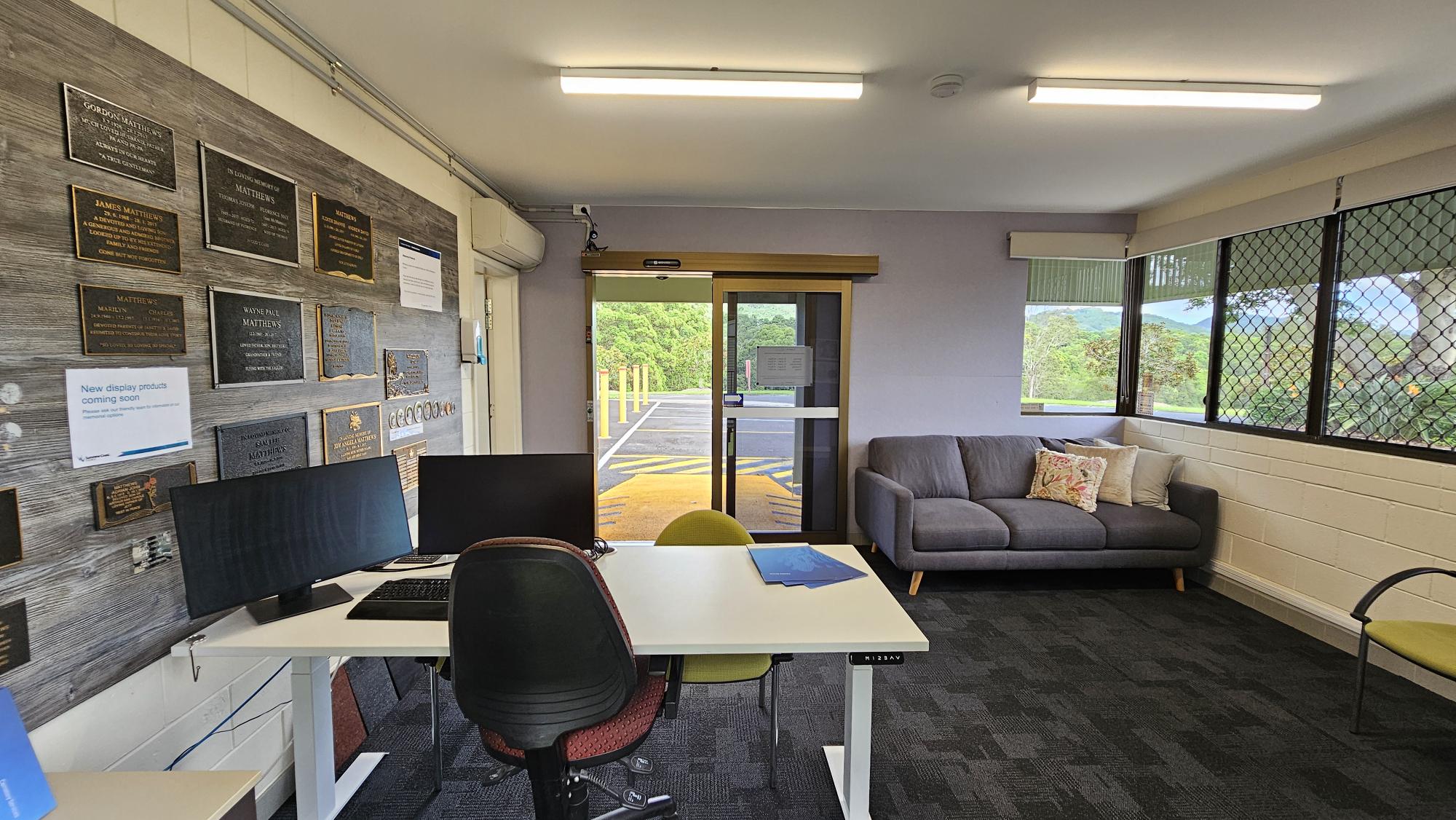 Office interior with a desk and two chairs in the foreground, featuring two computer monitors. The left wall displays numerous plaques. In the background, a glass door leads outside, and a grey sofa with cushions is positioned against the right wall. Large windows with mesh screens provide a view of greenery outside. The floor is carpeted in a dark pattern, and the ceiling has fluorescent lighting. Office interior with a desk and two chairs in the foreground, featuring two computer monitors. The left wall displays numerous plaques. In the background, a glass door leads outside, and a grey sofa with cushions is positioned against the right wall. Large windows with mesh screens provide a view of greenery outside. The floor is carpeted in a dark pattern, and the ceiling has fluorescent lighting.