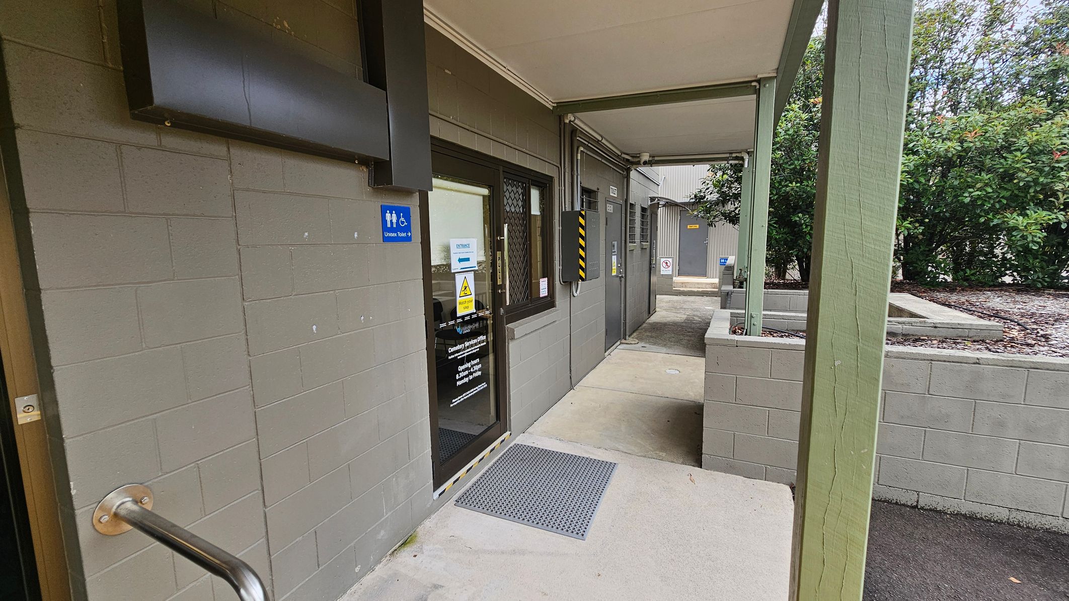 A covered walkway with a green-painted brick wall on the left, featuring a door with a glass panel and a sign indicating a universal toilet. A metal handrail is attached to the wall. The floor is concrete with a textured mat near the door. On the right, a green post supports the roof, and there are bushes in the background. Further along the walkway, there are additional doors and a small courtyard area. A covered walkway with a green-painted brick wall on the left, featuring a door with a glass panel and a sign indicating a universal toilet. A metal handrail is attached to the wall. The floor is concrete with a textured mat near the door. On the right, a green post supports the roof, and there are bushes in the background. Further along the walkway, there are additional doors and a small courtyard area.