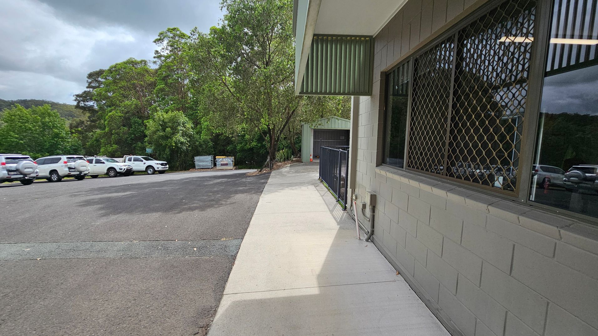 On the right, there's a building with a grey brick wall and a window covered by a metal grid. A narrow concrete path runs alongside the building. On the left, several white vehicles are parked on an asphalt surface. In the background, there are dense green trees and a couple of large bins. The sky is overcast, casting a muted light over the scene. On the right, there's a building with a grey brick wall and a window covered by a metal grid. A narrow concrete path runs alongside the building. On the left, several white vehicles are parked on an asphalt surface. In the background, there are dense green trees and a couple of large bins. The sky is overcast, casting a muted light over the scene.