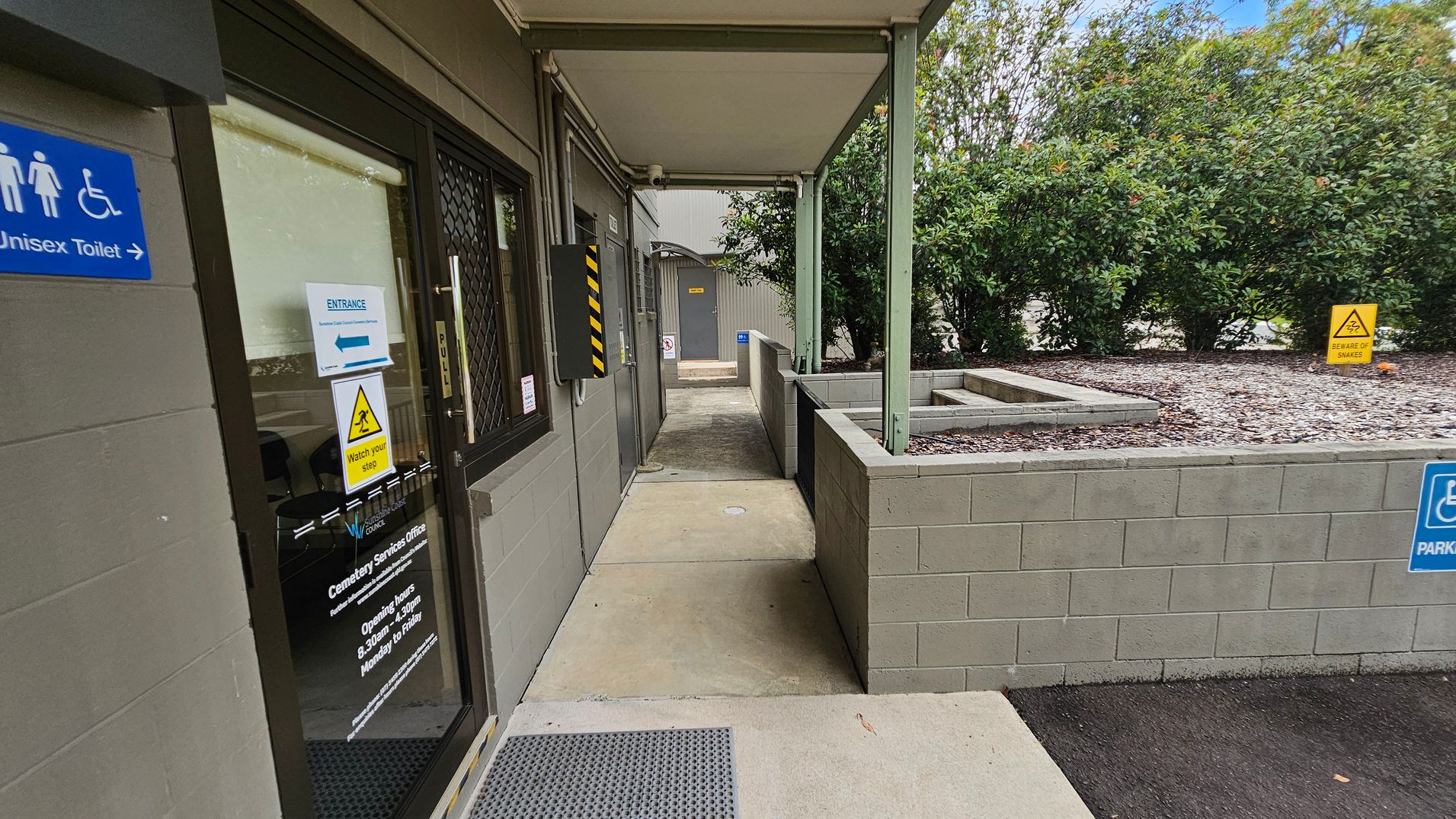 On the left, there is a glass door with signage indicating a unisex toilet and entrance information. The door has a "Watch your step" warning. In the middle, a covered walkway leads to another door in the distance. On the right, a low wall with a garden bed filled with gravel and a "Beware of snakes" sign. Bushes are visible in the background. The floor is concrete, and the ceiling of the walkway is flat and supported by green metal beams. On the left, there is a glass door with signage indicating a unisex toilet and entrance information. The door has a "Watch your step" warning. In the middle, a covered walkway leads to another door in the distance. On the right, a low wall with a garden bed filled with gravel and a "Beware of snakes" sign. Bushes are visible in the background. The floor is concrete, and the ceiling of the walkway is flat and supported by green metal beams.