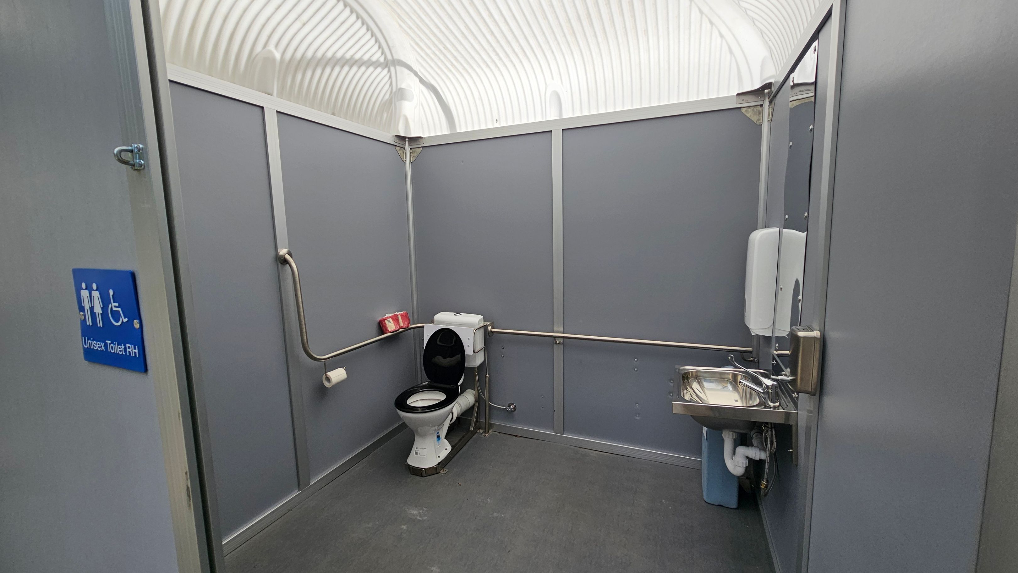 A grey-walled unisex toilet with accessibility features. On the left, a blue sign indicates "Unisex Toilet RH". In the middle, a toilet with a black seat is positioned against the wall, with a grab rail beside it. A roll of toilet paper is mounted on the wall. On the right, a stainless steel sink with a soap dispenser above it. The ceiling is arched and translucent, allowing light to filter through. The floor is grey and appears to be made of a non-slip material. A grey-walled unisex toilet with accessibility features. On the left, a blue sign indicates "Unisex Toilet RH". In the middle, a toilet with a black seat is positioned against the wall, with a grab rail beside it. A roll of toilet paper is mounted on the wall. On the right, a stainless steel sink with a soap dispenser above it. The ceiling is arched and translucent, allowing light to filter through. The floor is grey and appears to be made of a non-slip material.