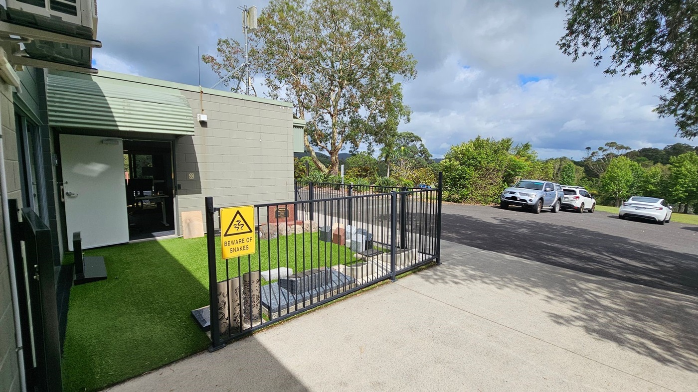 On the left, there's a building with a light green exterior and an open white door. A small area of artificial grass is enclosed by a black metal fence with a "Beware of Snakes" sign. In the middle, a tree is visible behind the building. On the right, a paved area with three parked cars is surrounded by greenery. The ground is a mix of concrete and asphalt. On the left, there's a building with a light green exterior and an open white door. A small area of artificial grass is enclosed by a black metal fence with a "Beware of Snakes" sign. In the middle, a tree is visible behind the building. On the right, a paved area with three parked cars is surrounded by greenery. The ground is a mix of concrete and asphalt.