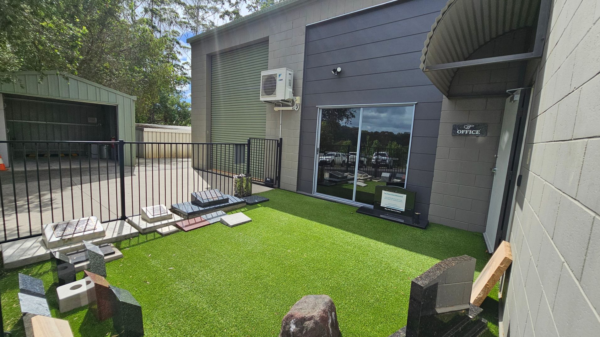 A small outdoor area with artificial grass on the ground. On the left, there is a metal fence with various stone samples displayed along its base. Behind the fence, a shed with an open door is visible. On the right, a building with a grey exterior features a sliding glass door and a sign marked "Office" next to it. An air conditioning unit is mounted above the door. Trees are visible in the background. A small outdoor area with artificial grass on the ground. On the left, there is a metal fence with various stone samples displayed along its base. Behind the fence, a shed with an open door is visible. On the right, a building with a grey exterior features a sliding glass door and a sign marked "Office" next to it. An air conditioning unit is mounted above the door. Trees are visible in the background.