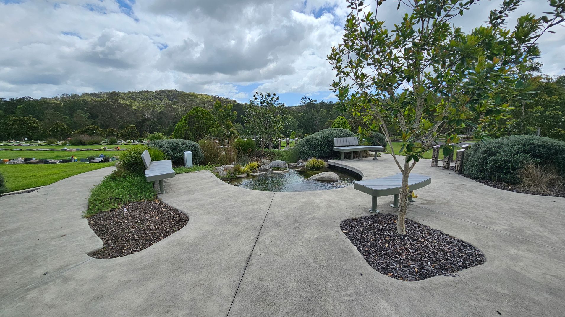 A landscaped garden with a concrete path winding through the centre. In the middle, there's a small tree surrounded by a circular patch of mulch. Benches are placed along the path, with shrubs and bushes neatly arranged around them. The background features a dense line of trees, and the grass is well-maintained. The sky is overcast with clouds. A landscaped garden with a concrete path winding through the centre. In the middle, there's a small tree surrounded by a circular patch of mulch. Benches are placed along the path, with shrubs and bushes neatly arranged around them. The background features a dense line of trees, and the grass is well-maintained. The sky is overcast with clouds.