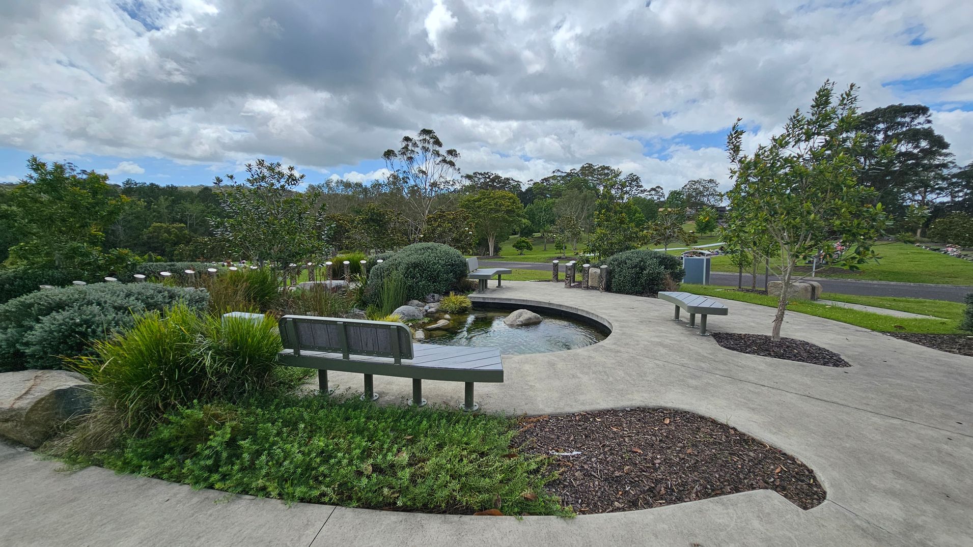 A landscaped garden with a curved concrete path and a small pond in the centre, surrounded by rocks and lush greenery. Wooden benches are placed along the path, offering seating. Trees and shrubs are scattered throughout the area, with a grassy field and more trees in the background. The sky is overcast with thick clouds. A landscaped garden with a curved concrete path and a small pond in the centre, surrounded by rocks and lush greenery. Wooden benches are placed along the path, offering seating. Trees and shrubs are scattered throughout the area, with a grassy field and more trees in the background. The sky is overcast with thick clouds.