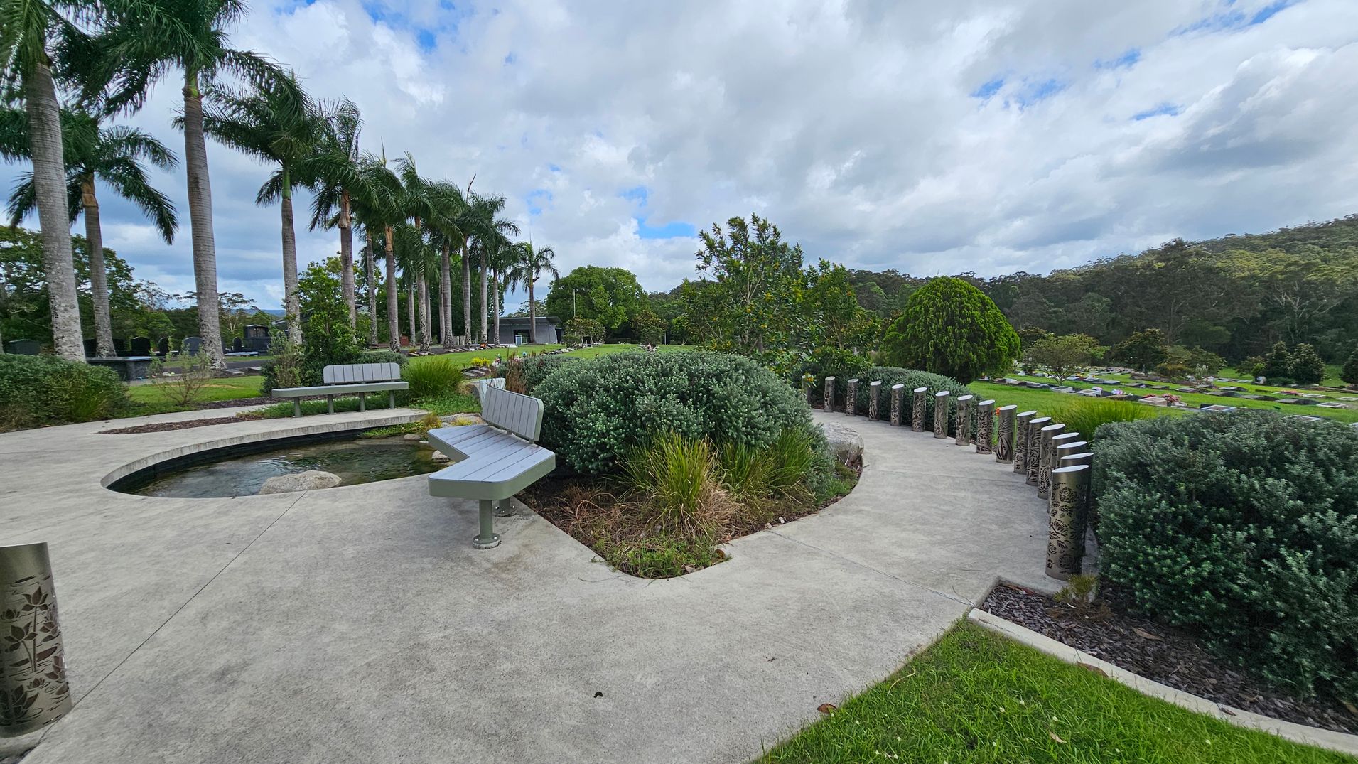 A landscaped garden with a curved concrete path and a small pond in the centre. On the left, there are tall palm trees lined up, and a few benches are placed along the path. Bushes and various plants are scattered throughout the area. On the right, decorative metal posts line the path, and more greenery extends into the distance. The background features a wooded area with dense trees. A landscaped garden with a curved concrete path and a small pond in the centre. On the left, there are tall palm trees lined up, and a few benches are placed along the path. Bushes and various plants are scattered throughout the area. On the right, decorative metal posts line the path, and more greenery extends into the distance. The background features a wooded area with dense trees.