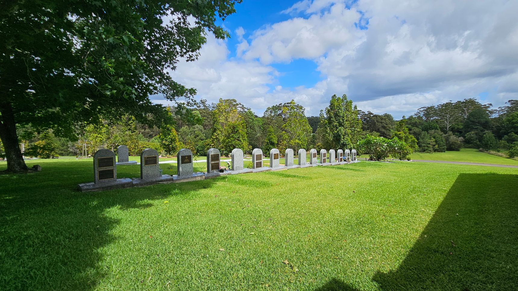 A row of gravestones is aligned on a well-maintained grassy area. On the right, there is a modern structure with a metal roof and open sides, featuring picnic tables underneath. In the background, lush green trees and a gently sloping landscape are visible. Overhead, large tree branches with dense foliage provide shade. A row of gravestones is aligned on a well-maintained grassy area. On the right, there is a modern structure with a metal roof and open sides, featuring picnic tables underneath. In the background, lush green trees and a gently sloping landscape are visible. Overhead, large tree branches with dense foliage provide shade.