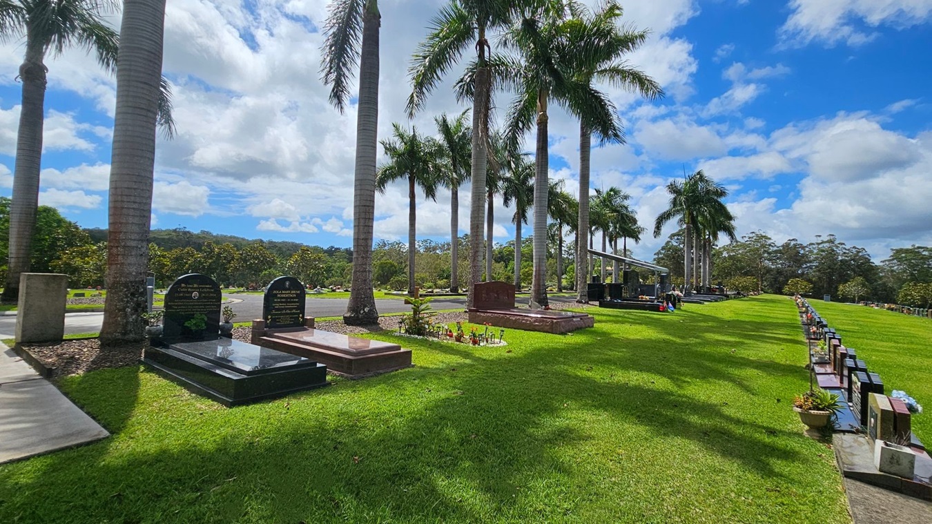 A cemetery with neatly arranged gravestones on the left and right. Tall palm trees line the area, casting shadows on the well-maintained green grass. In the background, there are more gravestones and a dense line of trees. The sky is partly cloudy, providing a serene atmosphere. A cemetery with neatly arranged gravestones on the left and right. Tall palm trees line the area, casting shadows on the well-maintained green grass. In the background, there are more gravestones and a dense line of trees. The sky is partly cloudy, providing a serene atmosphere.