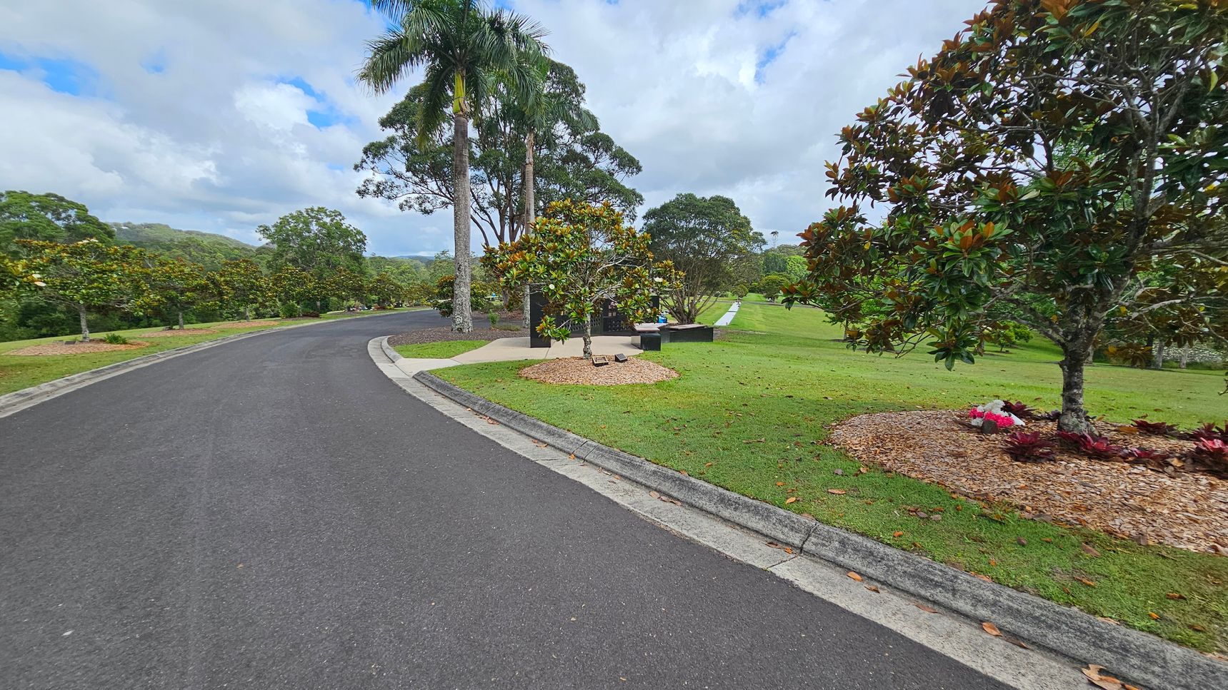 A winding asphalt road curves to the left, bordered by a grassy verge. On the right, a well-maintained lawn features several trees, including a large one with dense foliage and a small flower arrangement at its base. In the background, tall palm trees and other greenery are visible, with a bench and a paved area nearby. The scene is lush and verdant, suggesting a park or garden setting. A winding asphalt road curves to the left, bordered by a grassy verge. On the right, a well-maintained lawn features several trees, including a large one with dense foliage and a small flower arrangement at its base. In the background, tall palm trees and other greenery are visible, with a bench and a paved area nearby. The scene is lush and verdant, suggesting a park or garden setting.