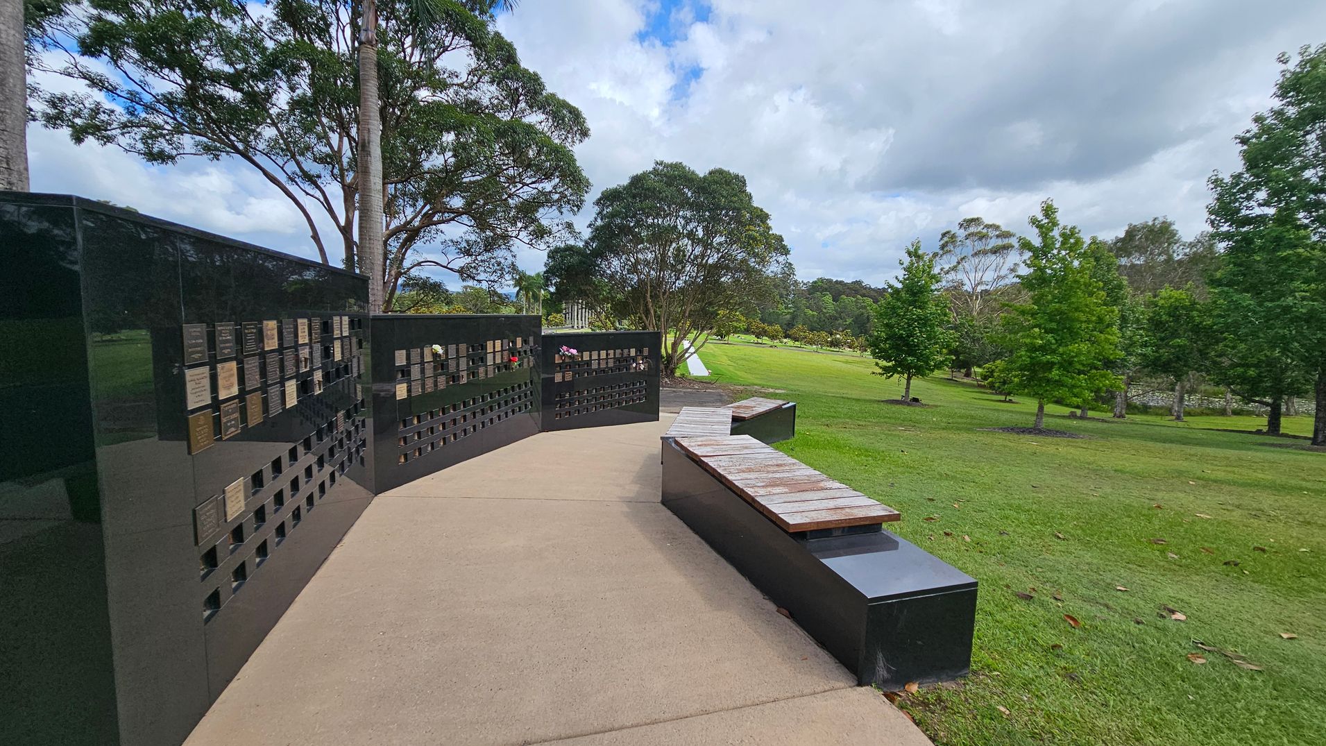 A paved pathway leads to a black memorial wall on the left, featuring numerous plaques. In front of the wall, there are wooden benches on black bases. The right side of the image shows a grassy area with scattered trees, including a large tree in the centre. The background is filled with more trees, creating a lush, green landscape. A paved pathway leads to a black memorial wall on the left, featuring numerous plaques. In front of the wall, there are wooden benches on black bases. The right side of the image shows a grassy area with scattered trees, including a large tree in the centre. The background is filled with more trees, creating a lush, green landscape.