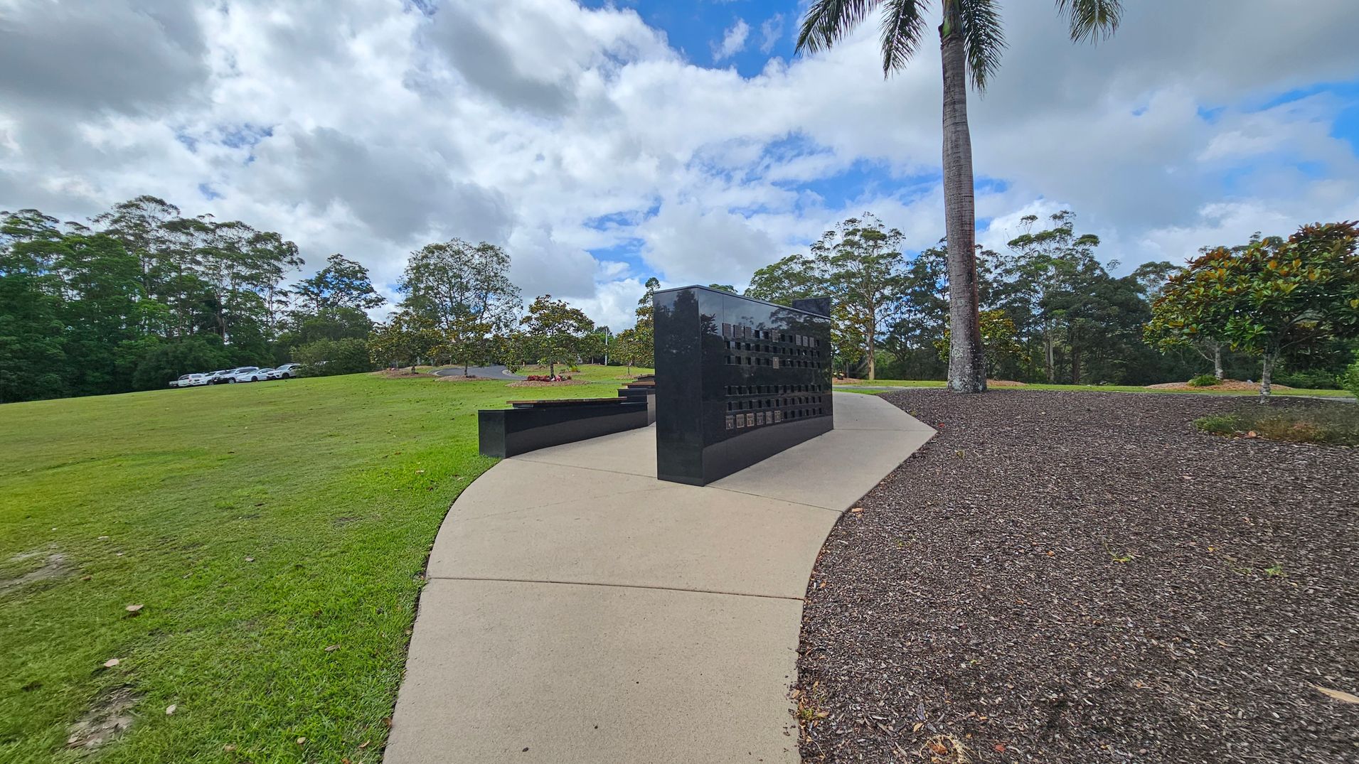 A black, polished memorial wall stands on a curved concrete path. To the left, there's a grassy area with several parked white cars in the distance. On the right, a tall palm tree and a small bush are situated on a bed of dark mulch. In the background, a line of trees borders the scene under a cloudy sky. A black, polished memorial wall stands on a curved concrete path. To the left, there's a grassy area with several parked white cars in the distance. On the right, a tall palm tree and a small bush are situated on a bed of dark mulch. In the background, a line of trees borders the scene under a cloudy sky.