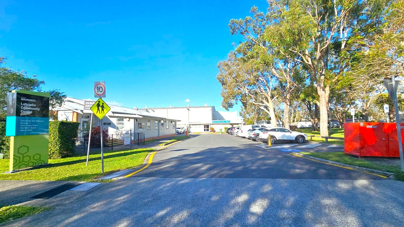 A paved road leads to a building in the centre, with a sign on the left reading "Labrador Community Centre." A speed limit sign and pedestrian crossing sign are nearby. On the right, several cars are parked under tall trees, and a red hydrant box is visible. The building on the left is single-storey with a light exterior. The area is surrounded by greenery, and the sky is clear and blue. A paved road leads to a building in the centre, with a sign on the left reading "Labrador Community Centre." A speed limit sign and pedestrian crossing sign are nearby. On the right, several cars are parked under tall trees, and a red hydrant box is visible. The building on the left is single-storey with a light exterior. The area is surrounded by greenery, and the sky is clear and blue.