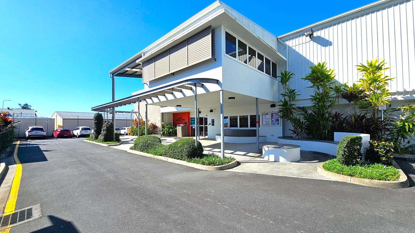 A modern building with a white exterior and large windows on the upper level. The entrance is sheltered by a metal awning supported by poles. To the right, there are landscaped areas with neatly trimmed bushes and tall plants. On the left, a driveway leads to a parking area with several cars. The ground is paved with asphalt, and the building's facade features a combination of horizontal and vertical lines. A modern building with a white exterior and large windows on the upper level. The entrance is sheltered by a metal awning supported by poles. To the right, there are landscaped areas with neatly trimmed bushes and tall plants. On the left, a driveway leads to a parking area with several cars. The ground is paved with asphalt, and the building's facade features a combination of horizontal and vertical lines.