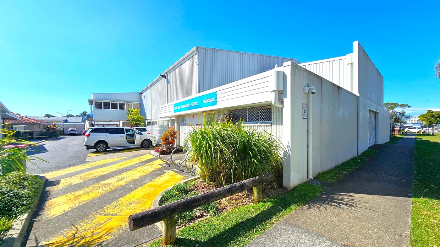A white building with a blue sign reading "Labrador Community Centre" is on the right. In front, there are green plants and a bicycle rack. A white vehicle is parked on a yellow-striped area to the left. The background shows more buildings and a few parked cars. The ground is a mix of concrete and grass, and a wooden barrier separates the path from the grass. The sky is clear and blue. A white building with a blue sign reading "Labrador Community Centre" is on the right. In front, there are green plants and a bicycle rack. A white vehicle is parked on a yellow-striped area to the left. The background shows more buildings and a few parked cars. The ground is a mix of concrete and grass, and a wooden barrier separates the path from the grass. The sky is clear and blue.
