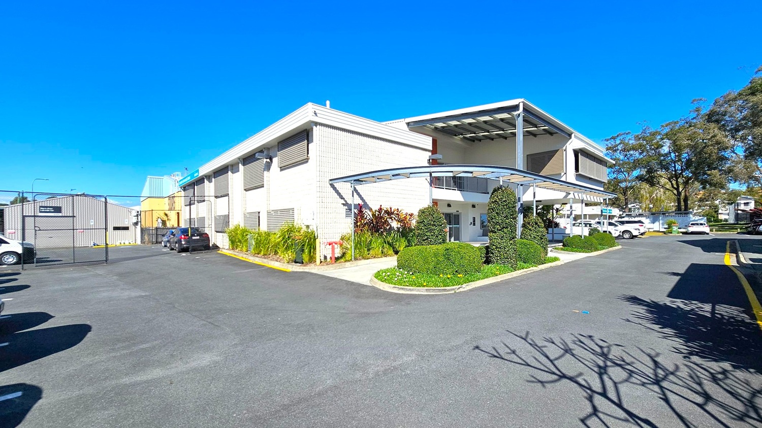 A modern building with a light grey facade and large windows is on the right. It features a covered entrance with neatly trimmed hedges and plants. To the left, a gated area with parked vehicles and an industrial-style building is visible. The foreground shows a wide asphalt driveway with shadows from nearby trees. In the background, more trees and parked cars are visible, adding to the suburban setting. A modern building with a light grey facade and large windows is on the right. It features a covered entrance with neatly trimmed hedges and plants. To the left, a gated area with parked vehicles and an industrial-style building is visible. The foreground shows a wide asphalt driveway with shadows from nearby trees. In the background, more trees and parked cars are visible, adding to the suburban setting.