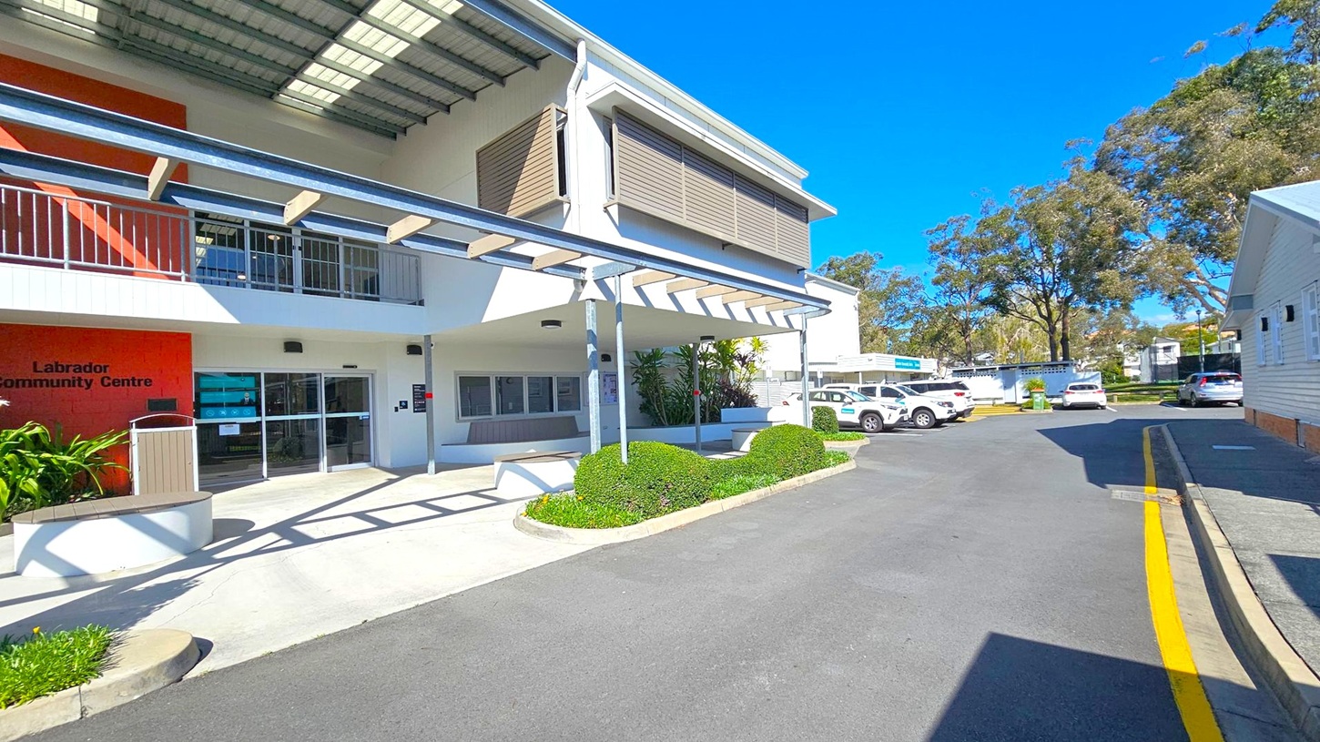 A two-storey building with a modern design, featuring a covered entrance and a sign reading "Labrador Community Centre" on the left. The building has large windows and a metal awning. In the foreground, there are neatly trimmed bushes and a paved walkway. On the right, several parked cars are visible along a driveway, with trees and additional buildings in the background. The sky is clear and blue. A two-storey building with a modern design, featuring a covered entrance and a sign reading "Labrador Community Centre" on the left. The building has large windows and a metal awning. In the foreground, there are neatly trimmed bushes and a paved walkway. On the right, several parked cars are visible along a driveway, with trees and additional buildings in the background. The sky is clear and blue.