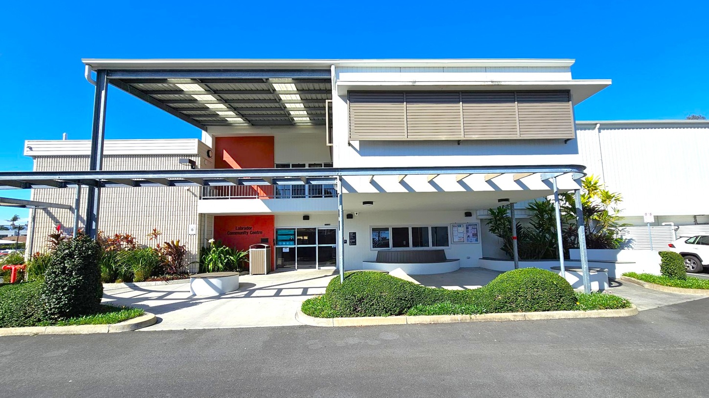 A modern building with a flat roof and a large overhang. The facade is a mix of white and orange, with a prominent entrance featuring glass doors. A metal awning extends over the walkway, casting shadows on the ground. Well-maintained shrubs and plants line the front, with a small garden area on the left. A white car is partially visible on the right. The pavement is dark grey, contrasting with the light-coloured building. A modern building with a flat roof and a large overhang. The facade is a mix of white and orange, with a prominent entrance featuring glass doors. A metal awning extends over the walkway, casting shadows on the ground. Well-maintained shrubs and plants line the front, with a small garden area on the left. A white car is partially visible on the right. The pavement is dark grey, contrasting with the light-coloured building.