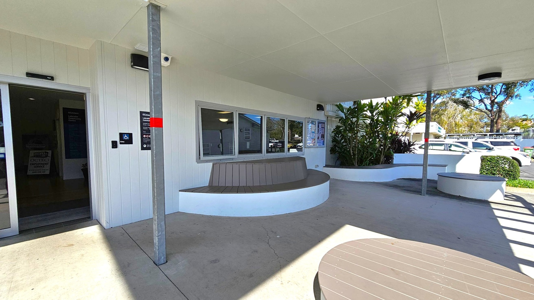 A covered outdoor area with a white ceiling and concrete floor. On the left, there's a dark entrance door with a sign indicating accessibility. In the centre, a curved bench is built into a white wall beneath a window. To the right, a planter with green plants and a small bench is visible. A metal pole supports the ceiling. In the background, several parked cars and trees are visible. A round table is partially visible in the foreground. A covered outdoor area with a white ceiling and concrete floor. On the left, there's a dark entrance door with a sign indicating accessibility. In the centre, a curved bench is built into a white wall beneath a window. To the right, a planter with green plants and a small bench is visible. A metal pole supports the ceiling. In the background, several parked cars and trees are visible. A round table is partially visible in the foreground.