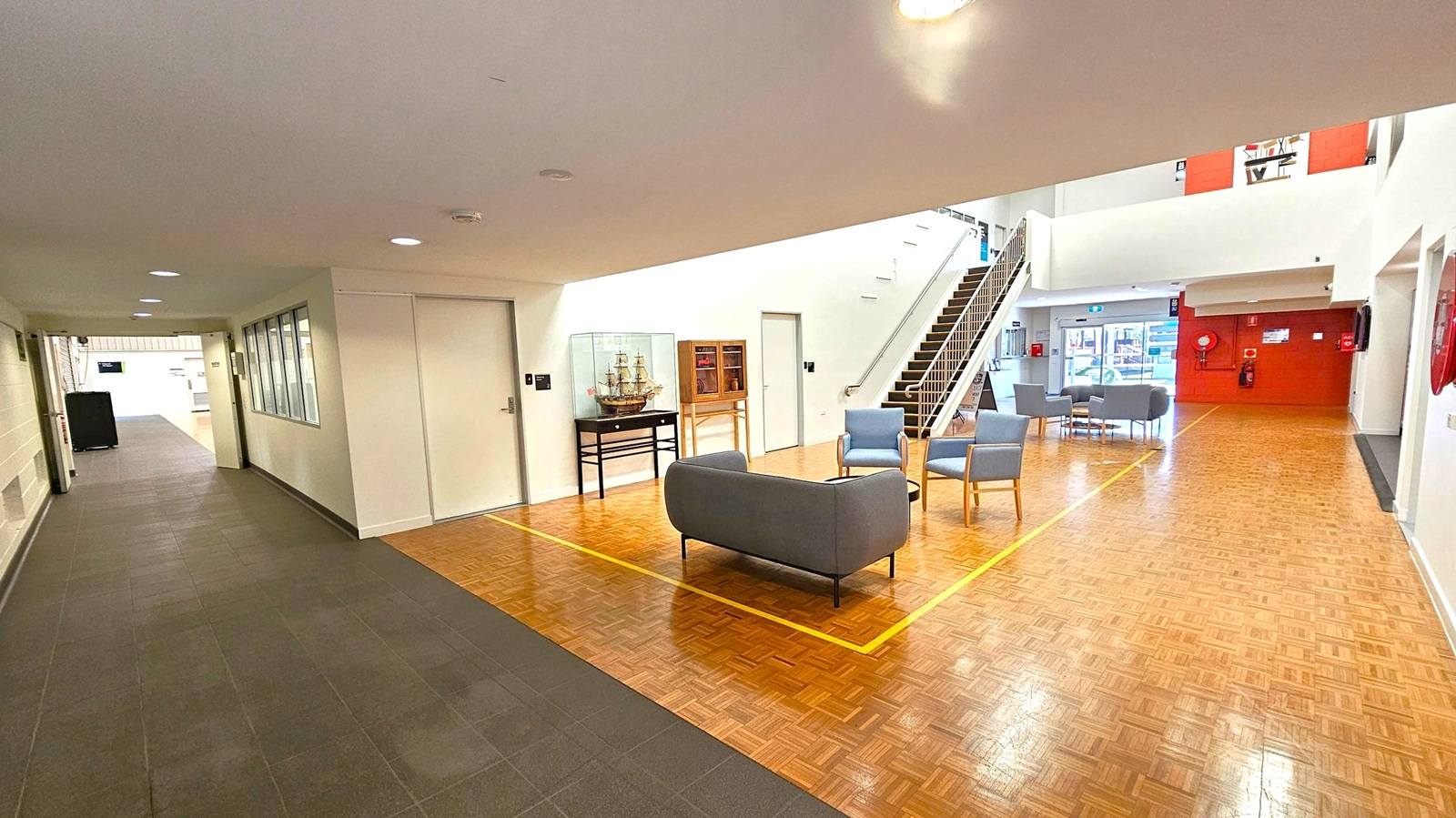 A spacious indoor area with a parquet floor. On the left, a corridor with dark tiles and a row of windows. In the centre, a seating area with a grey sofa and blue chairs around a small table. A model ship in a glass case and a wooden cabinet are against the wall. A staircase with a metal railing leads to an upper level. On the right, a red wall with fire safety equipment. The ceiling is white with recessed lighting. A spacious indoor area with a parquet floor. On the left, a corridor with dark tiles and a row of windows. In the centre, a seating area with a grey sofa and blue chairs around a small table. A model ship in a glass case and a wooden cabinet are against the wall. A staircase with a metal railing leads to an upper level. On the right, a red wall with fire safety equipment. The ceiling is white with recessed lighting.