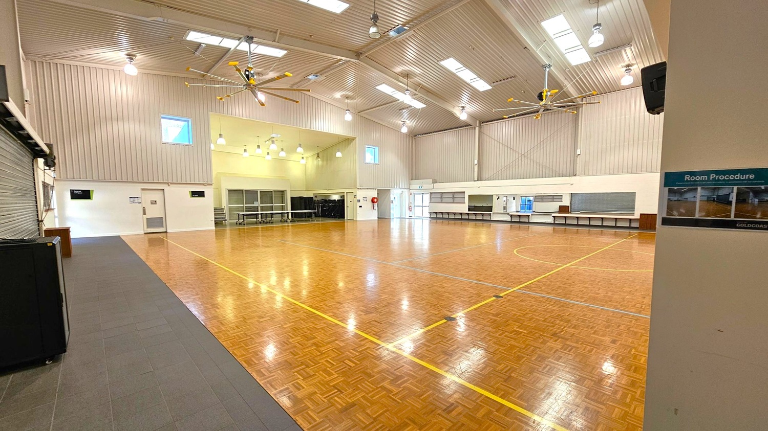 A large indoor hall with a polished wooden floor marked with yellow lines. The ceiling is high with skylights and large fans. On the left, there is a closed roller door and a small door with a sign above it. The right side has a long counter with a shutter and a sign that reads "Room Procedure." The back wall features windows and a fire extinguisher. The lighting consists of multiple hanging lamps. A large indoor hall with a polished wooden floor marked with yellow lines. The ceiling is high with skylights and large fans. On the left, there is a closed roller door and a small door with a sign above it. The right side has a long counter with a shutter and a sign that reads "Room Procedure." The back wall features windows and a fire extinguisher. The lighting consists of multiple hanging lamps.