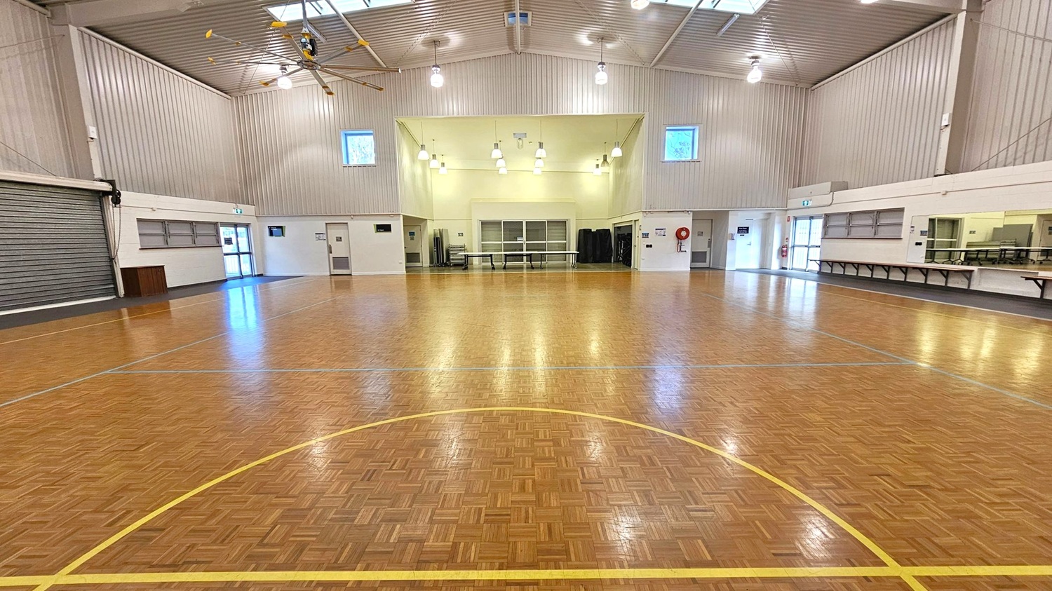 A spacious indoor hall with a polished wooden floor marked with yellow lines. The ceiling is high with exposed beams and several hanging lights. On the left, there is a large roller door and a small entrance. The back wall features two small windows and a central area with tables and stacked chairs. On the right, there are long tables and a serving area with a counter and windows. The walls are light-coloured, and the space is well-lit. A spacious indoor hall with a polished wooden floor marked with yellow lines. The ceiling is high with exposed beams and several hanging lights. On the left, there is a large roller door and a small entrance. The back wall features two small windows and a central area with tables and stacked chairs. On the right, there are long tables and a serving area with a counter and windows. The walls are light-coloured, and the space is well-lit.
