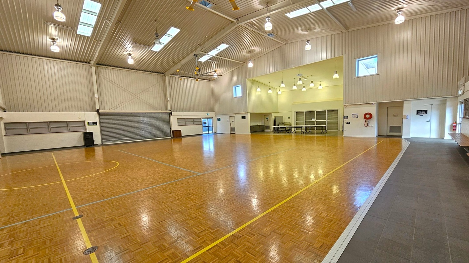 A spacious indoor sports hall with a polished wooden floor marked with yellow and blue lines. The ceiling is high with exposed beams and several skylights, along with hanging lights. On the left, there is a large roller door and a small exit door. The right side features a fire hose reel and a couple of doors, one with a sign. The back wall has a few windows and a section with tables and chairs. The floor transitions to a grey tiled area on the right. A spacious indoor sports hall with a polished wooden floor marked with yellow and blue lines. The ceiling is high with exposed beams and several skylights, along with hanging lights. On the left, there is a large roller door and a small exit door. The right side features a fire hose reel and a couple of doors, one with a sign. The back wall has a few windows and a section with tables and chairs. The floor transitions to a grey tiled area on the right.