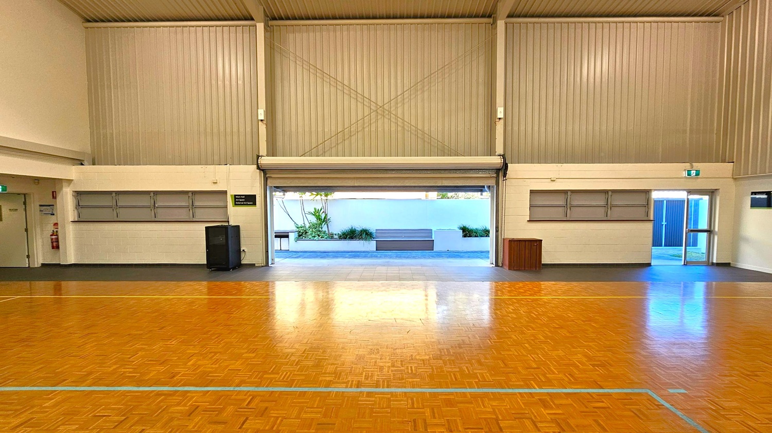 A large indoor space with a polished wooden floor and high corrugated metal walls. In the centre, a wide roller door is partially open, revealing an outdoor area with a white wall, plants, and a bench. On the left, there is a closed door and a fire extinguisher. On the right, a glass door leads outside. The ceiling is high and ribbed, and there are two sets of horizontal windows on either side of the roller door. A large indoor space with a polished wooden floor and high corrugated metal walls. In the centre, a wide roller door is partially open, revealing an outdoor area with a white wall, plants, and a bench. On the left, there is a closed door and a fire extinguisher. On the right, a glass door leads outside. The ceiling is high and ribbed, and there are two sets of horizontal windows on either side of the roller door.