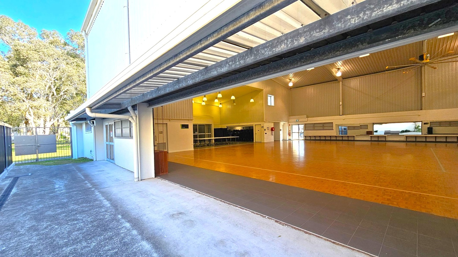 A large indoor space with a wooden floor and high ceiling, featuring multiple hanging lights and a large ceiling fan. The room is open to the outside through a wide, raised garage-style door. Outside, there is a concrete path leading to a metal gate, with trees visible in the background. The interior walls are light-coloured, and there are several windows and a door on the far wall. Tables are lined up against the wall inside. A large indoor space with a wooden floor and high ceiling, featuring multiple hanging lights and a large ceiling fan. The room is open to the outside through a wide, raised garage-style door. Outside, there is a concrete path leading to a metal gate, with trees visible in the background. The interior walls are light-coloured, and there are several windows and a door on the far wall. Tables are lined up against the wall inside.