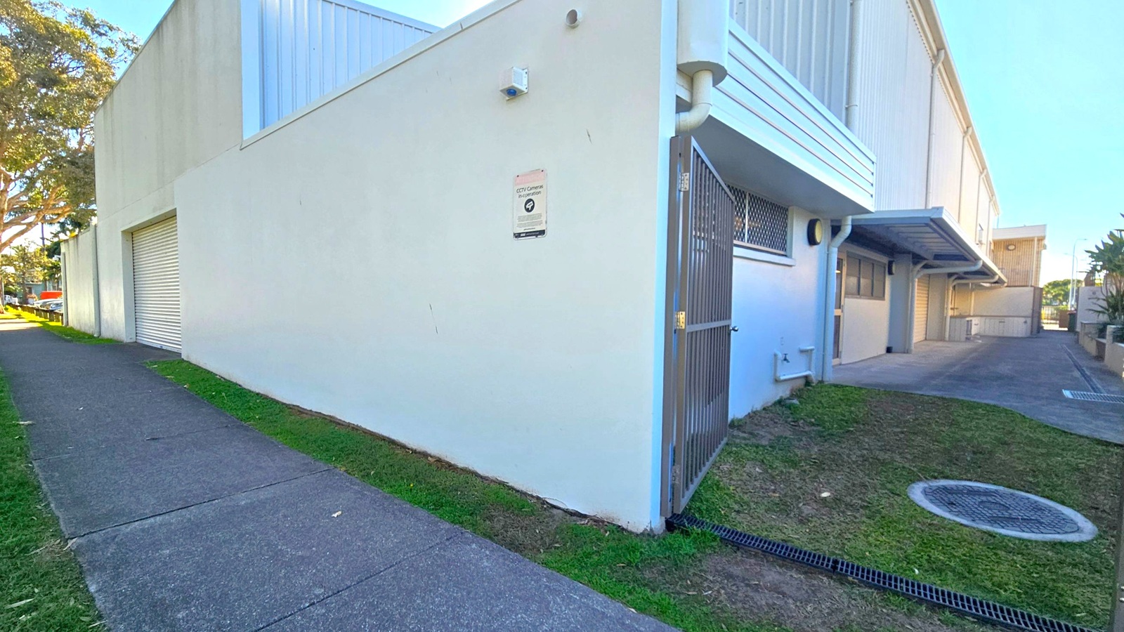 A large, light-coloured building with a flat roof and a roller door on the left side. A CCTV sign is mounted on the wall near a metal gate. The building has a small window with a security grille. A concrete path runs alongside the building, bordered by grass and a few trees. On the right, there is a narrow driveway leading to the back of the building, with a covered area and additional doors. The ground is a mix of concrete and grass. A large, light-coloured building with a flat roof and a roller door on the left side. A CCTV sign is mounted on the wall near a metal gate. The building has a small window with a security grille. A concrete path runs alongside the building, bordered by grass and a few trees. On the right, there is a narrow driveway leading to the back of the building, with a covered area and additional doors. The ground is a mix of concrete and grass.