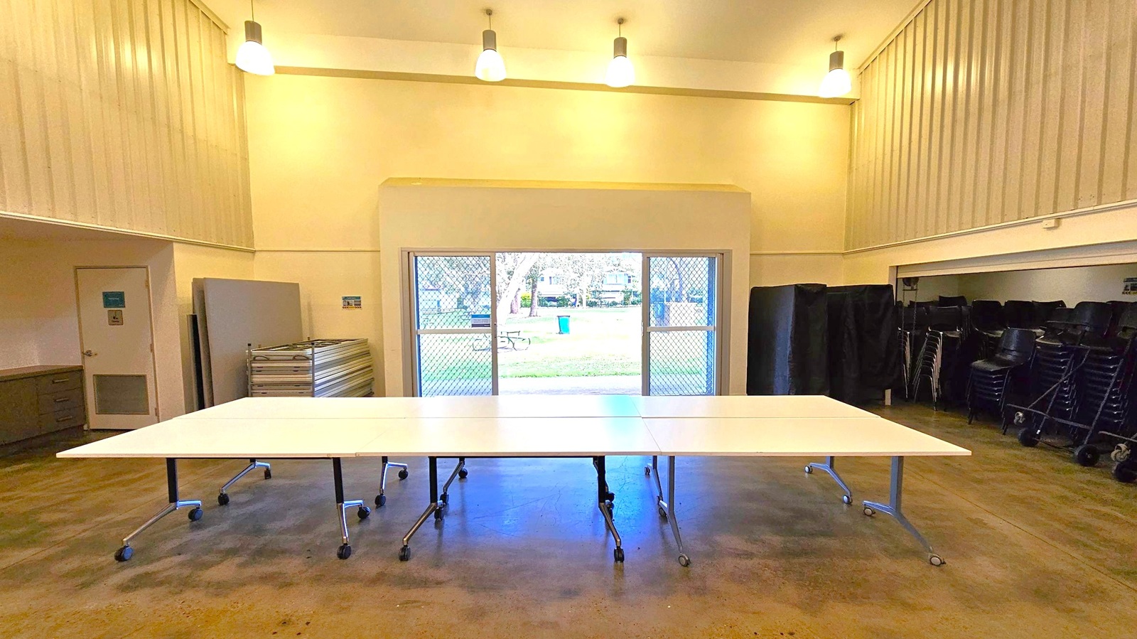 A room with a high ceiling and three pendant lights. In the centre, there are several white tables on wheels arranged together. The floor is a polished concrete surface. On the left, there is a stack of folded tables and a door. On the right, there are stacked chairs. The back wall features a large window with a mesh screen, offering a view of a grassy area with trees, a picnic table, and a blue bin. A room with a high ceiling and three pendant lights. In the centre, there are several white tables on wheels arranged together. The floor is a polished concrete surface. On the left, there is a stack of folded tables and a door. On the right, there are stacked chairs. The back wall features a large window with a mesh screen, offering a view of a grassy area with trees, a picnic table, and a blue bin.